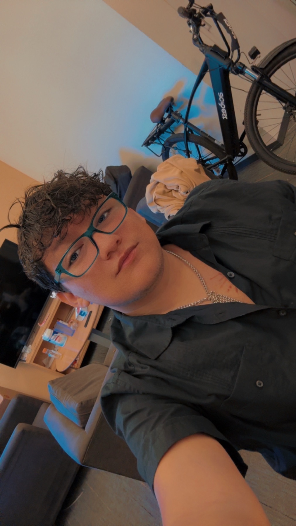 A man with curly hair and glasses takes a selfie while wearing a dark shirt. A bicycle is visible in the background.