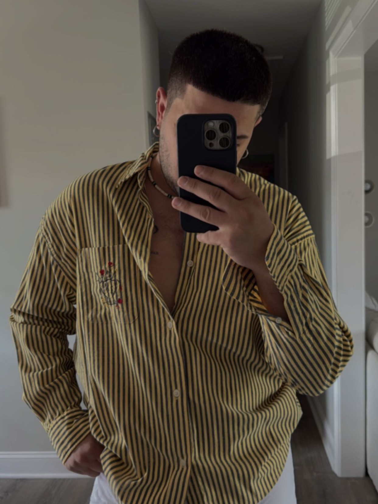 A man takes a mirror selfie, showcasing his outfit. He wears a yellow and black striped shirt, partially unbuttoned.