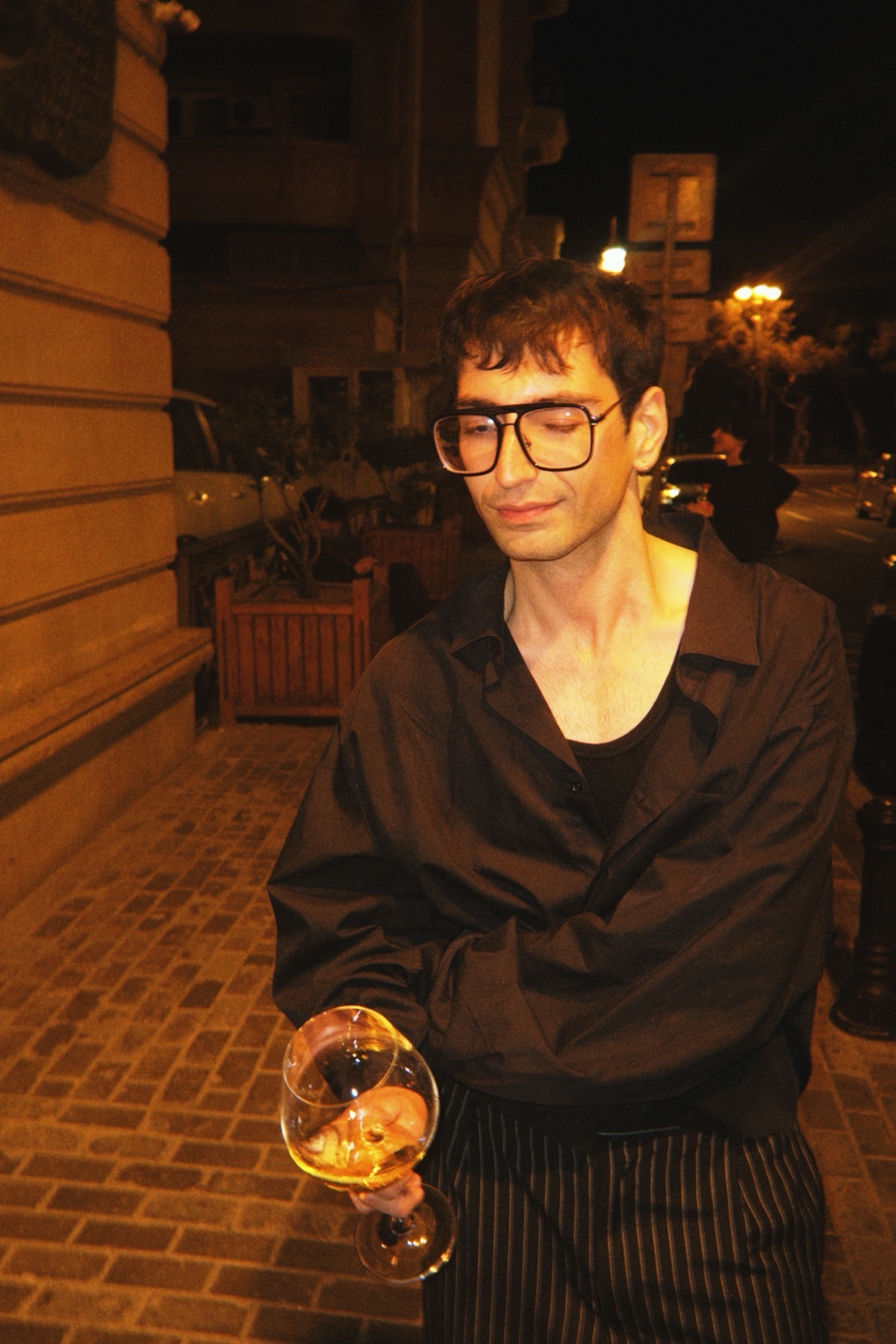 A man wearing large-framed glasses and a black button-down shirt over a tank top holds a wine glass in his left hand while standing on a brick-paved sidewalk at night.