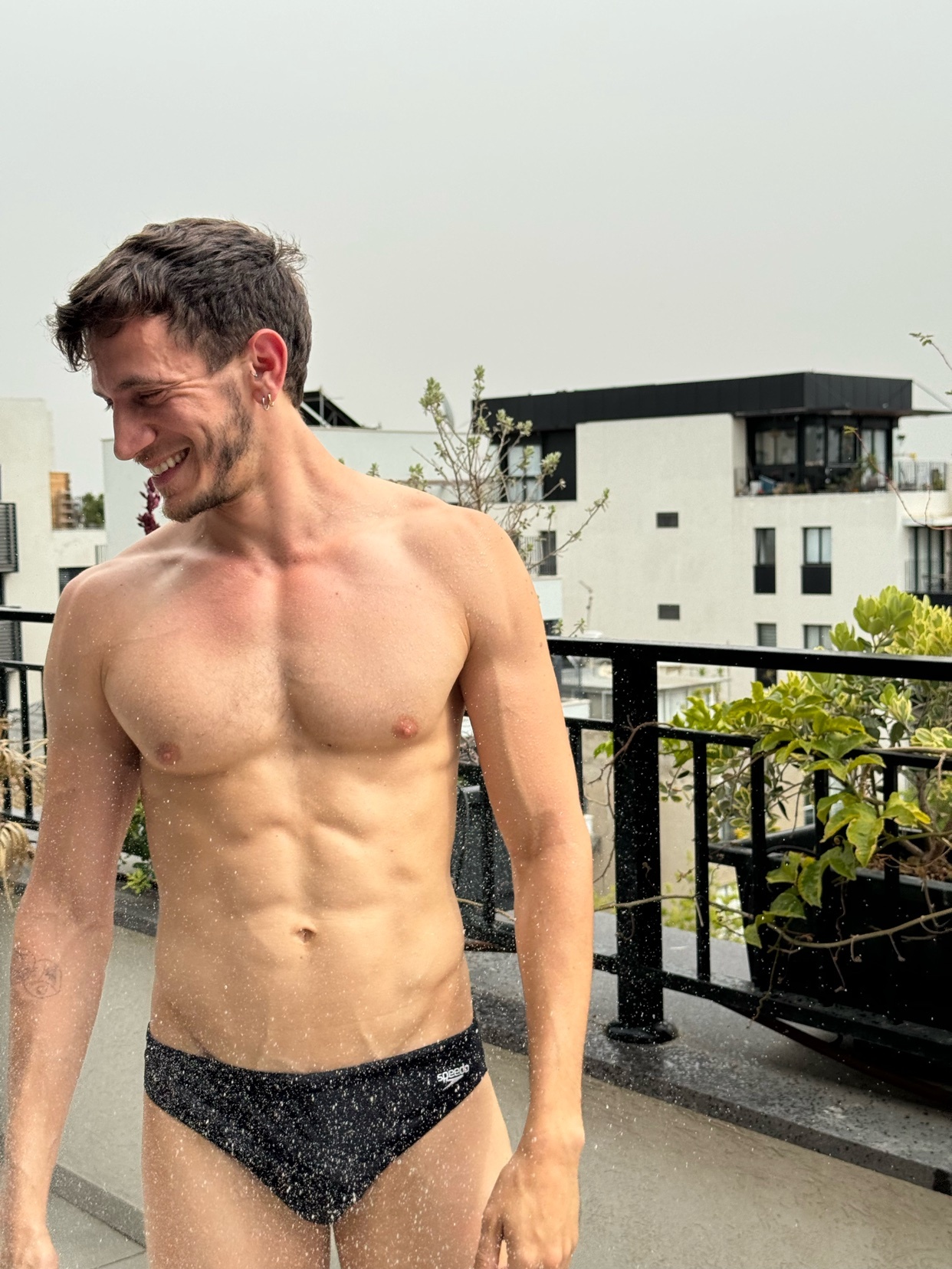 A shirtless man smiles while standing outside. He is wearing a swimsuit and has a muscular physique.