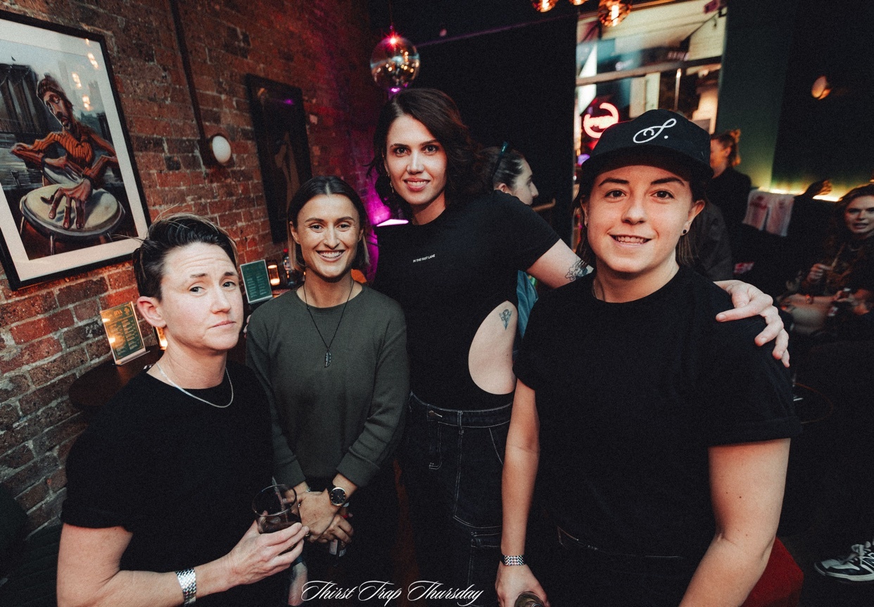 A group of four friends pose for a photo in a dimly lit bar. The two individuals on the right are wearing black t-shirts, one with a baseball cap. The two individuals on the left are also dressed casually. A disco ball is visible in the background.