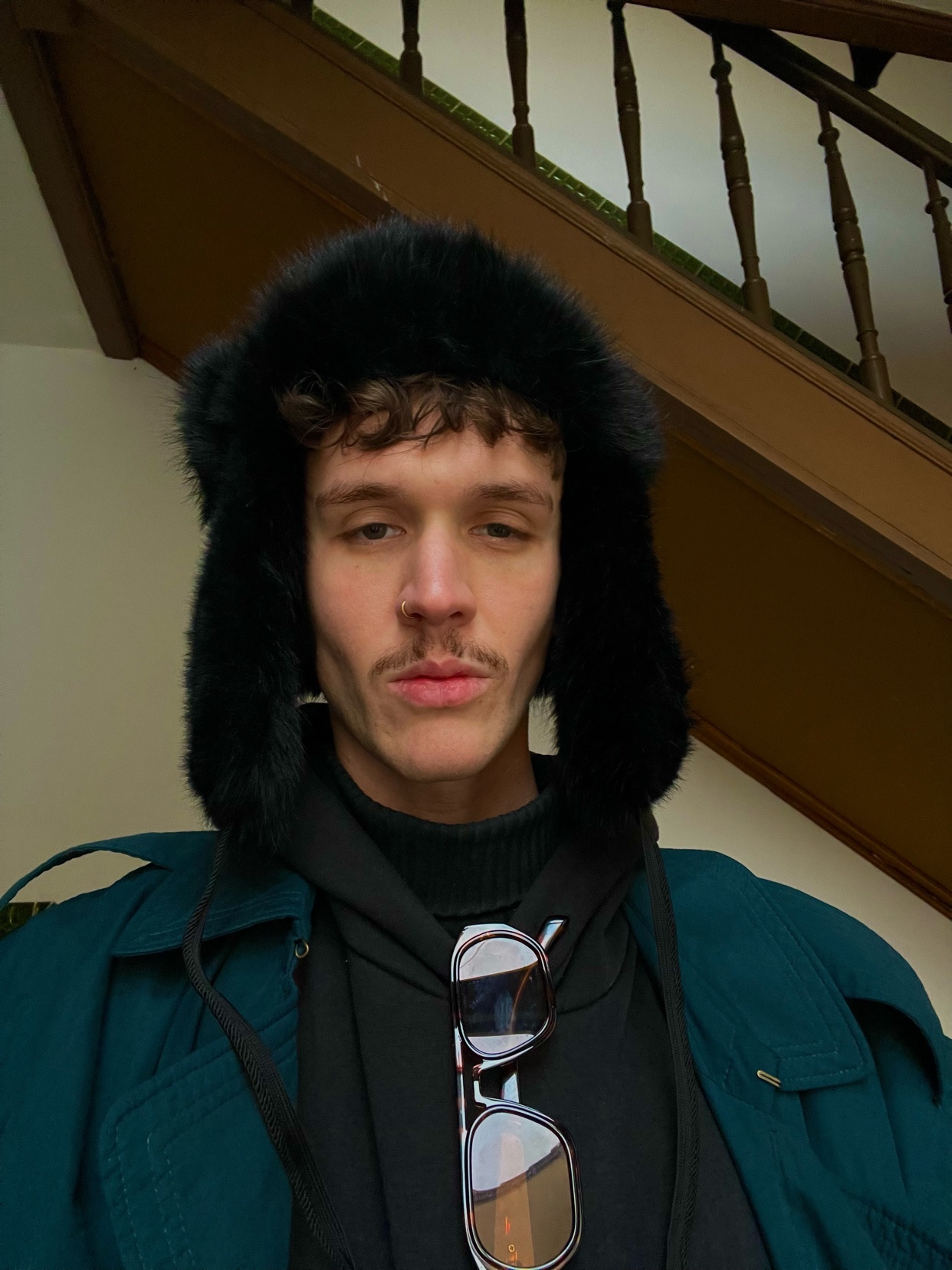A man poses for a selfie, wearing a stylish winter hat and coat. He has curly hair and a mustache.
