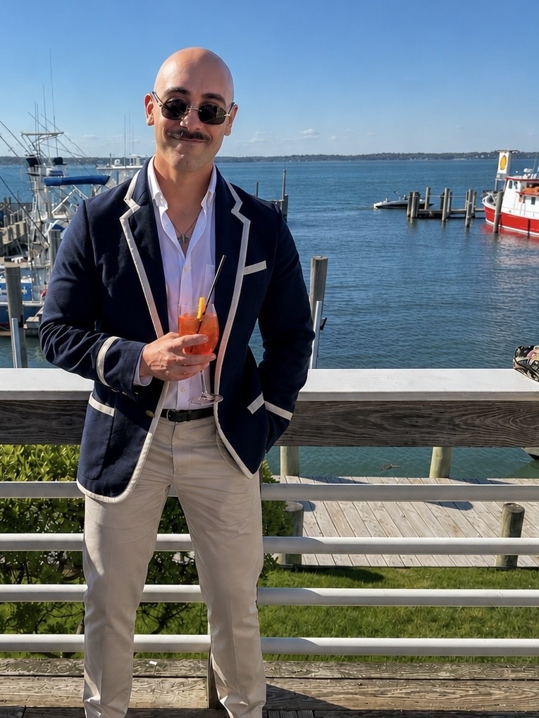 A bald man with a mustache and sunglasses poses for a photo while holding a cocktail. He is wearing a navy blazer with white trim, a white shirt, and tan pants. The background shows a body of water with boats and a pier.