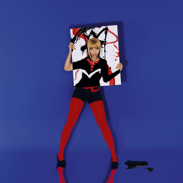 A woman poses with a piece of art in front of her face. She is wearing red tights, black shorts, and black heels.