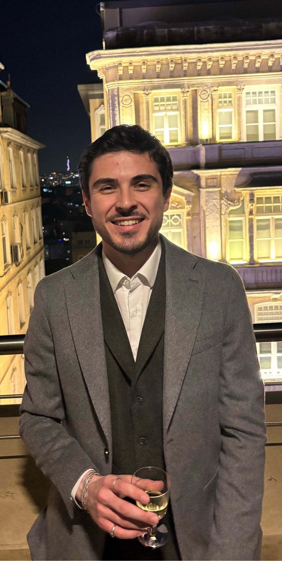 A man in a suit smiles while holding a glass of wine. The photo is taken at night with a cityscape in the background.