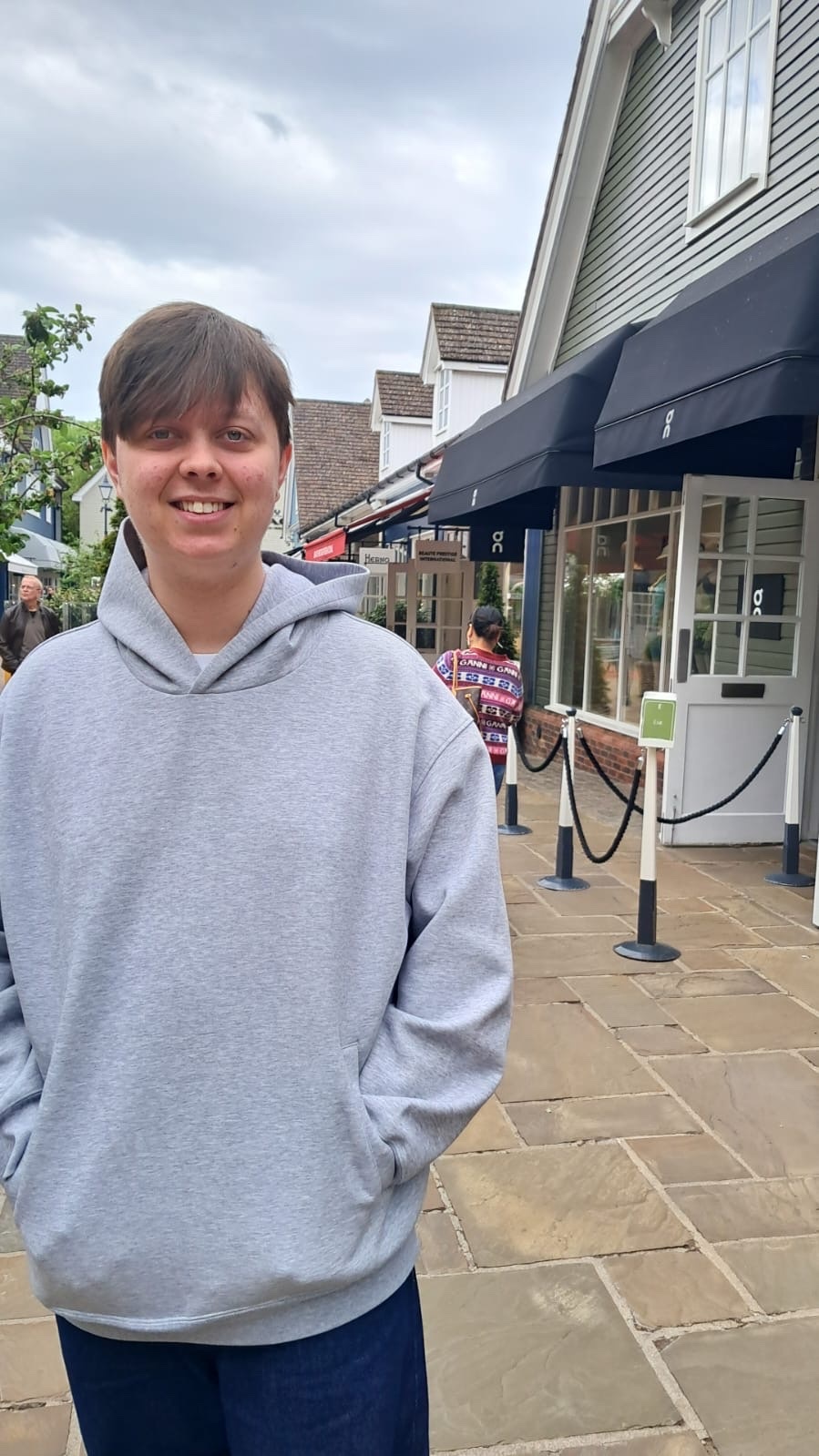 A non-binary person with short hair smiles at the camera while standing outside a row of shops. They are wearing a grey hoodie and dark pants.