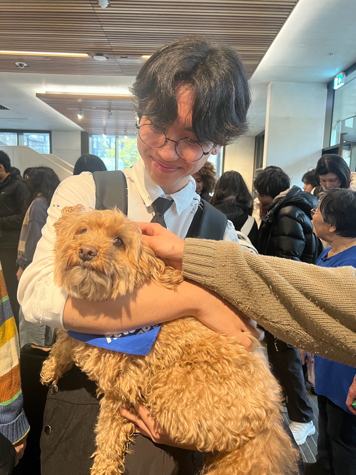 A young Asian man with glasses and dark, wavy hair smiles as he holds a fluffy, light brown dog wearing a blue bandana. The dog looks directly at the camera with a slightly concerned expression.