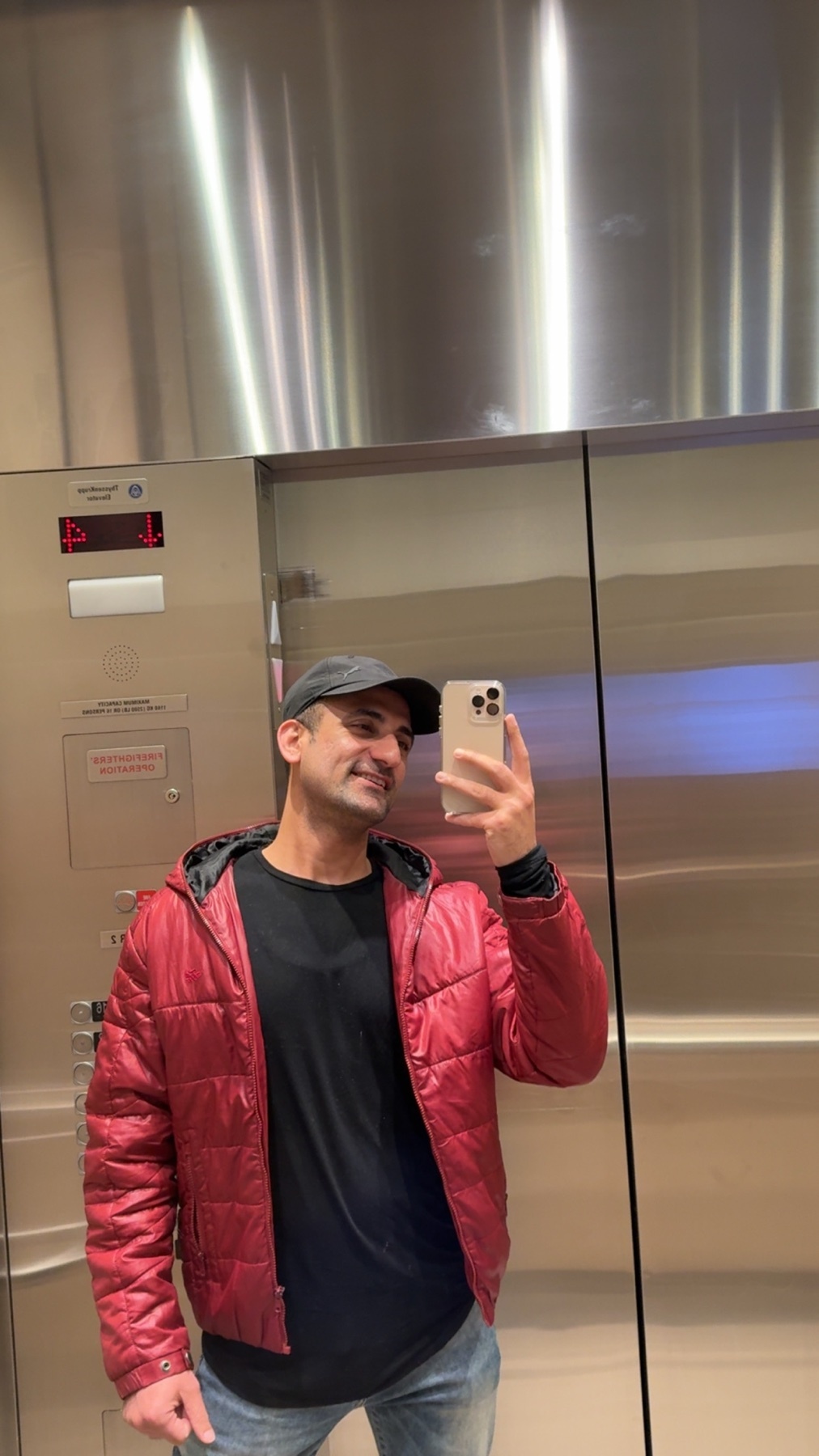 A man in a red puffy jacket and a black baseball cap takes a selfie in an elevator.