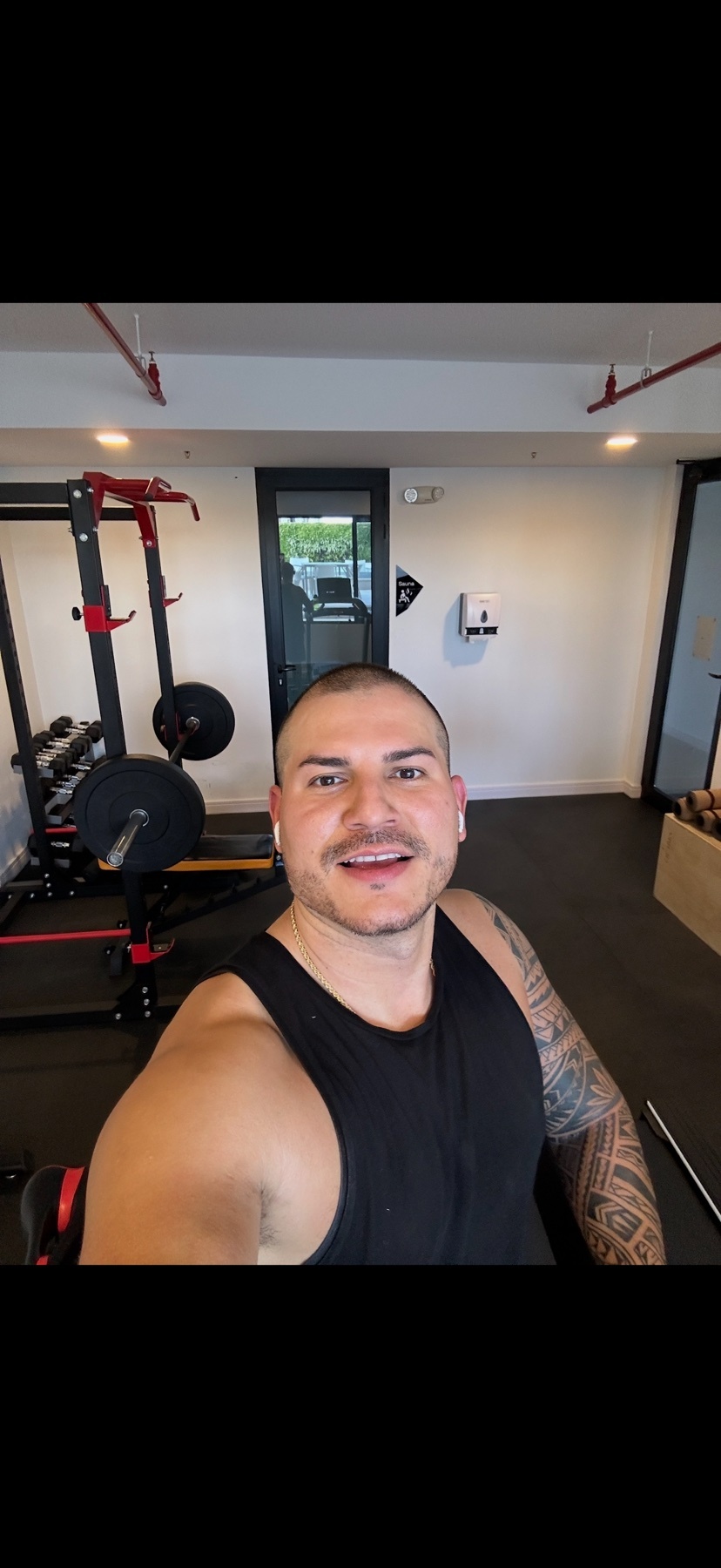 A man with tattoos and earbuds takes a selfie in a gym. He is wearing a black tank top and is smiling at the camera. Behind him, a weight rack with weights is visible.