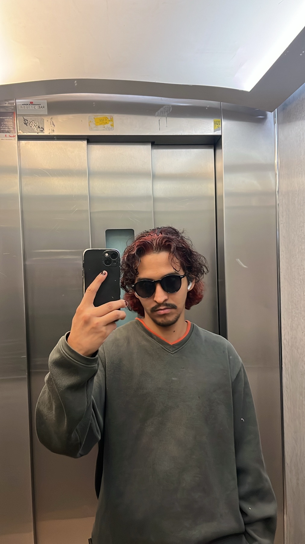 A man with curly, dark red hair and sunglasses takes a selfie in an elevator. He is wearing a dark grey sweatshirt and holding a black smartphone.