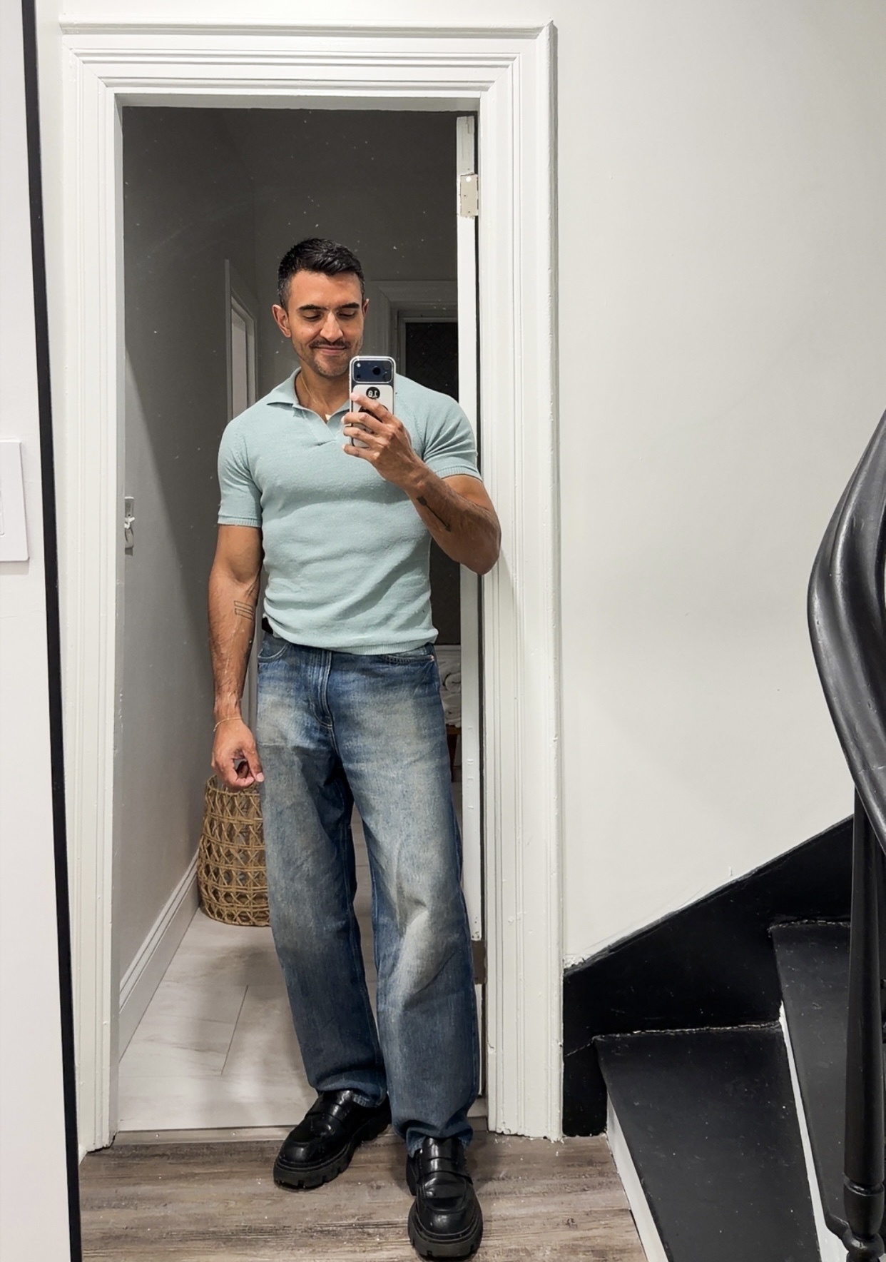 A man takes a mirror selfie in a doorway. He is wearing a light blue polo shirt and loose-fitting jeans.