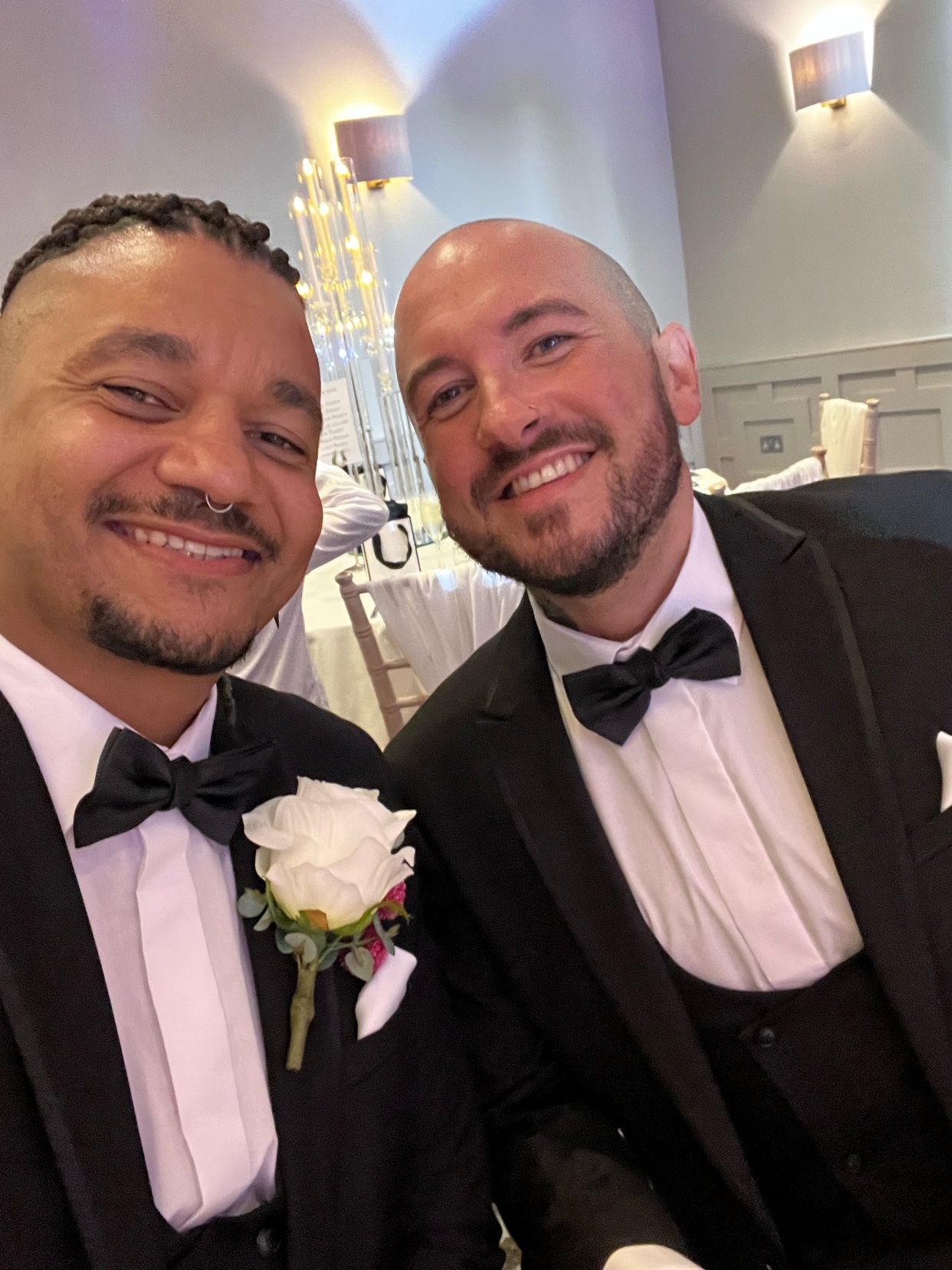 Two men in formal attire smile for a selfie. They appear to be at a wedding, with one wearing a boutonniere.