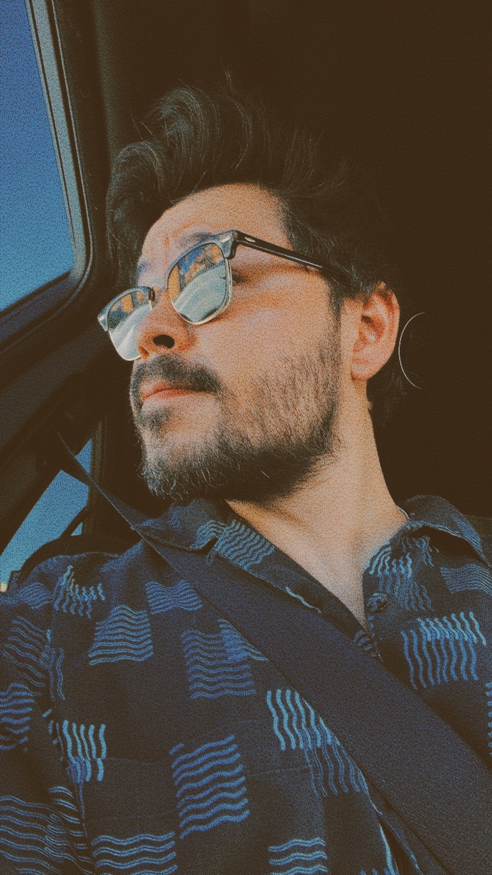 A man with a beard and stubble wears stylish glasses and a patterned blue shirt while looking out of a car window. The image has a vintage filter applied.