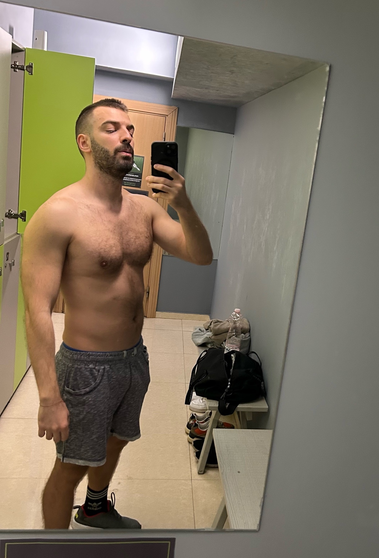 A man takes a mirror selfie in a gym locker room. He is shirtless, showing off his muscular physique.
