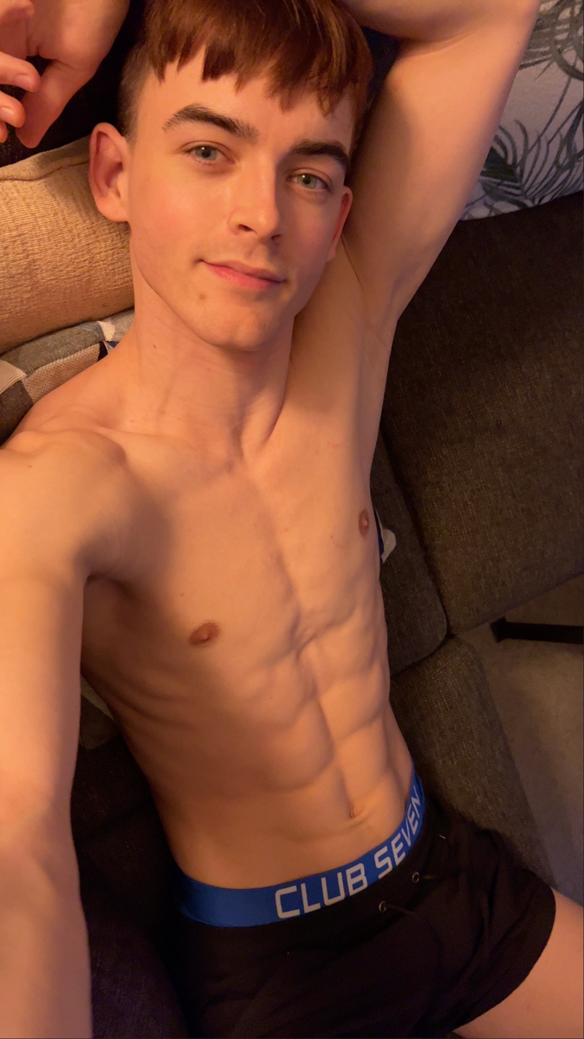 A man lies on a couch, looking at the camera. He is wearing black Club Seven thong underwear and has a toned physique with visible abs.