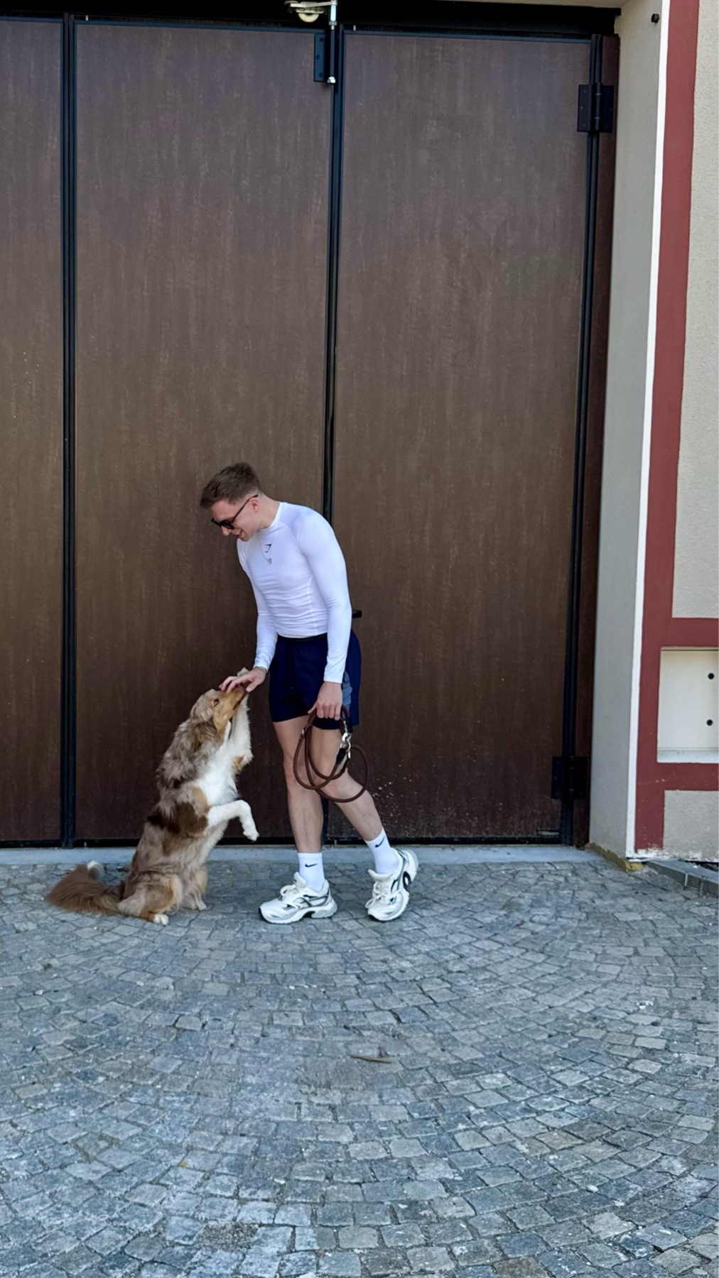 A man in activewear pets his Australian Shepherd dog while standing on a cobblestone path in front of a large wooden gate.