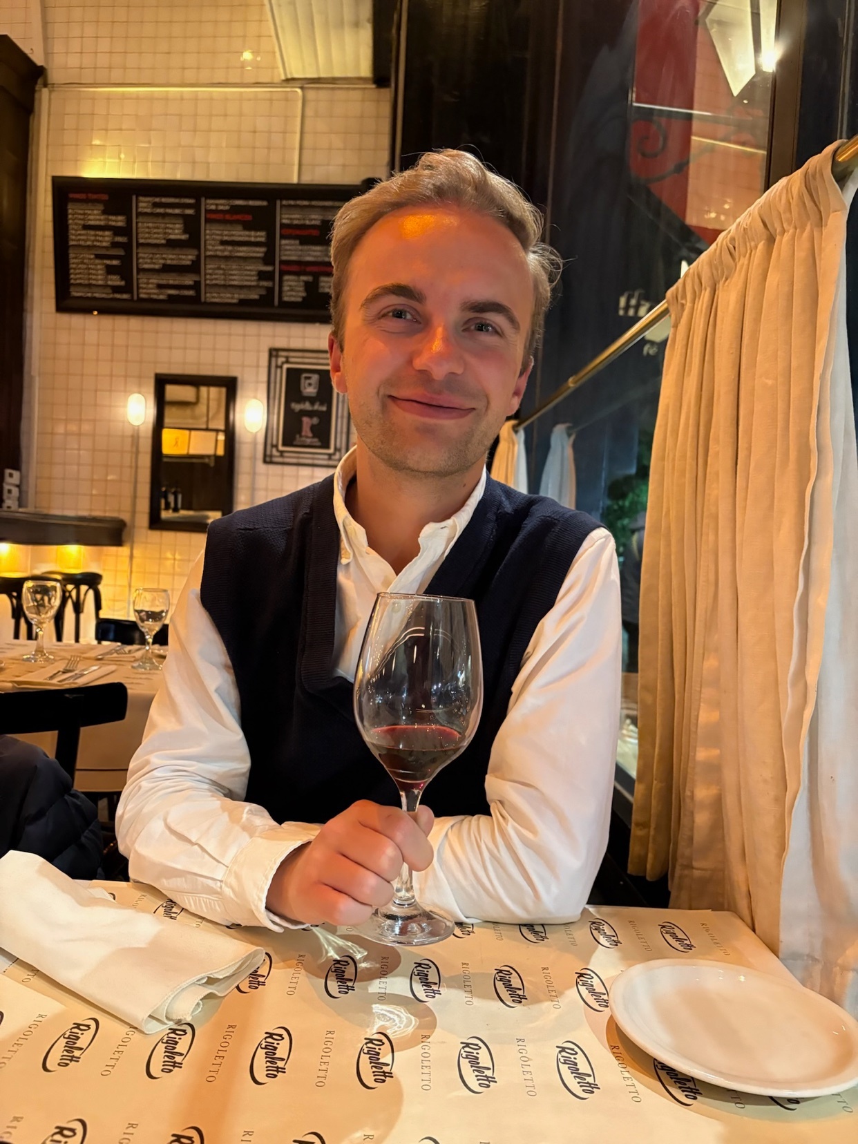 A man smiles while holding a glass of red wine at a table in a restaurant. The man is wearing a white shirt and a dark vest.
