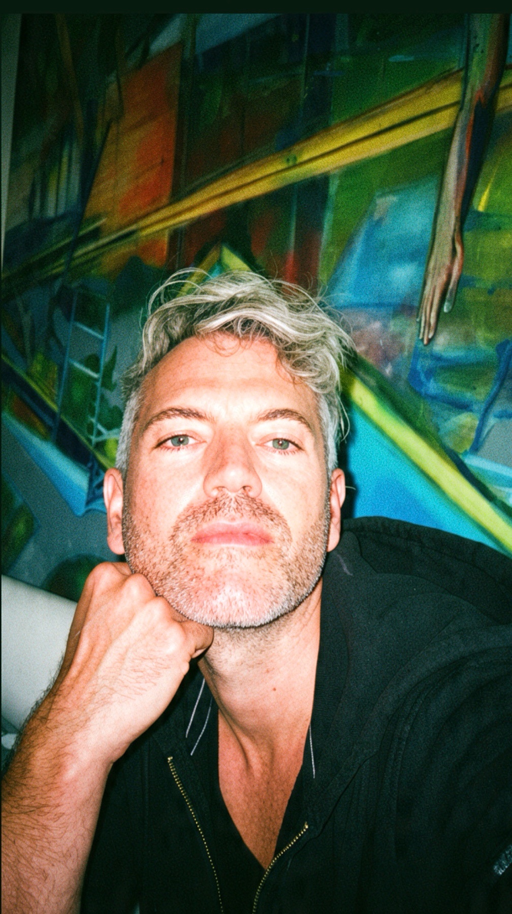 A man with grey hair and stubble rests his chin on his hand, looking at the camera. Behind him is a colorful, abstract painting.