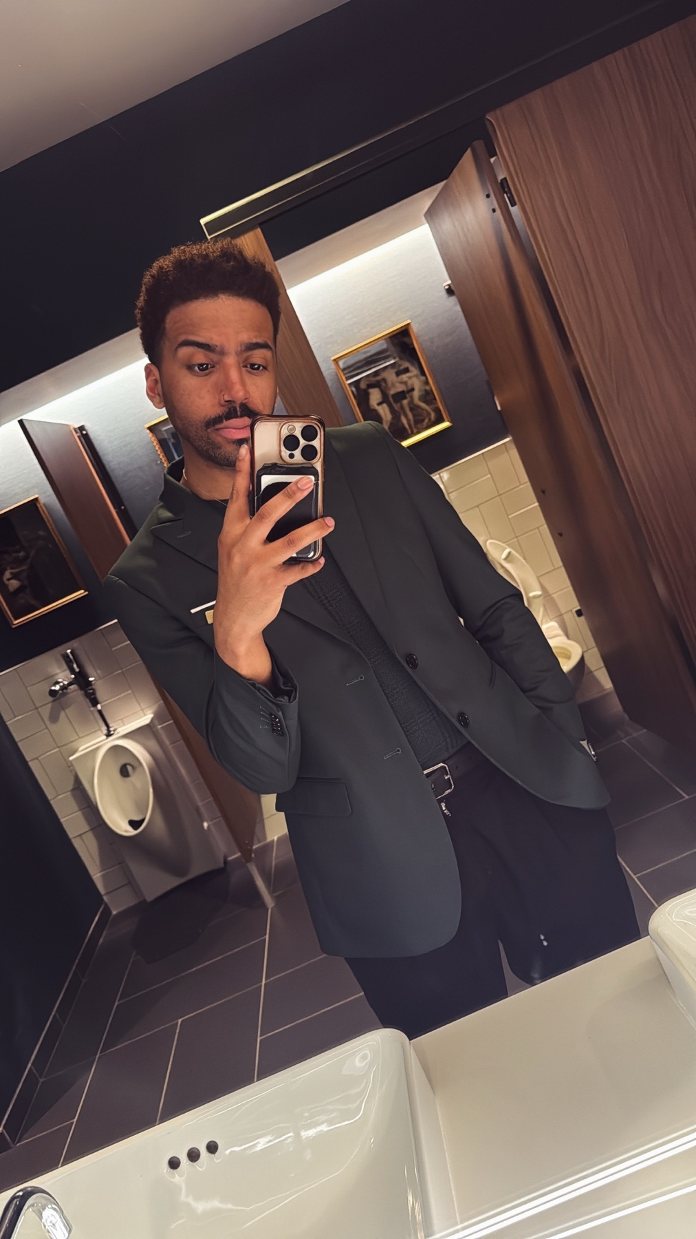 A man takes a mirror selfie in a stylish bathroom. He is wearing a dark green suit and has curly hair.
