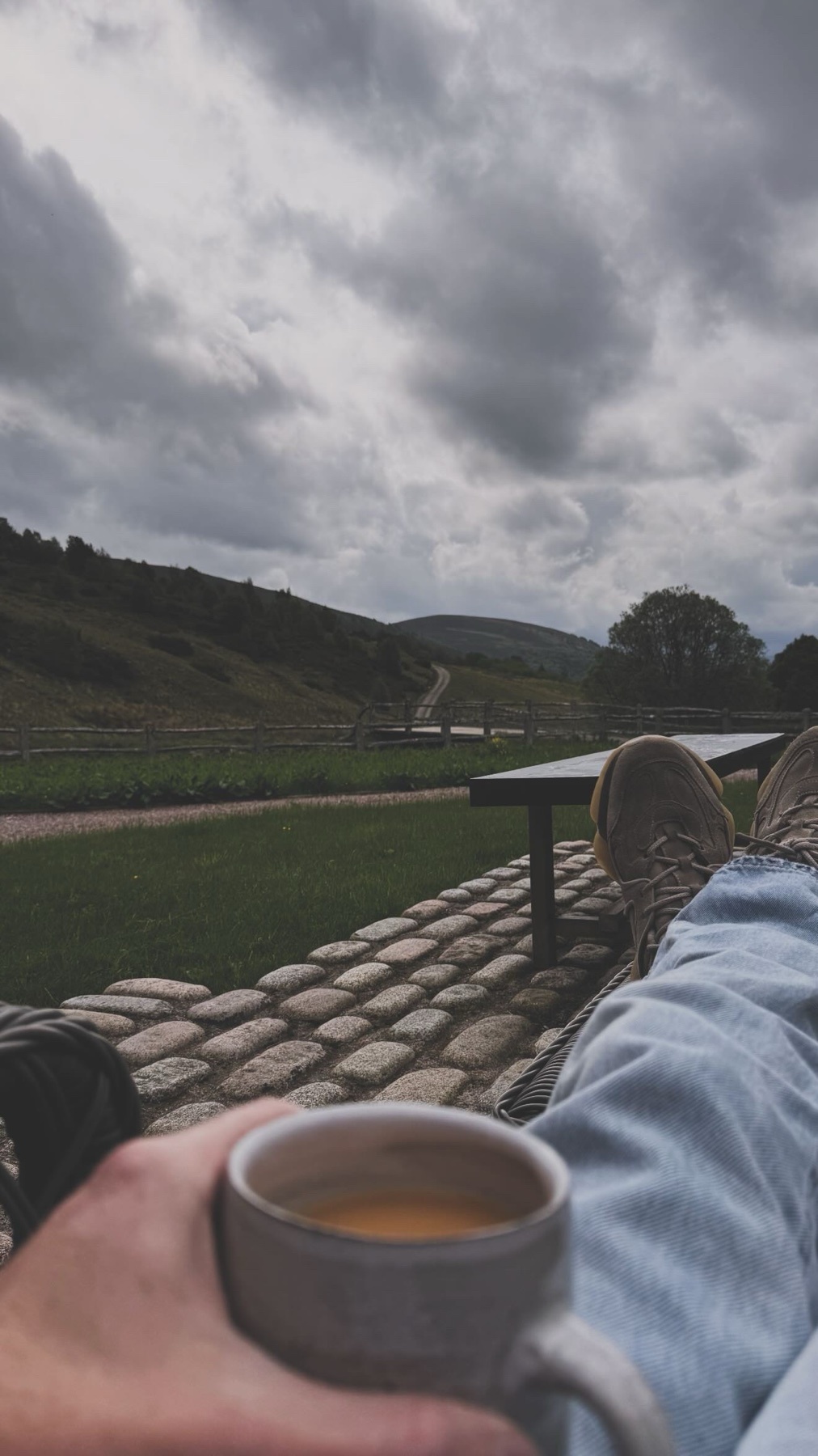 A person holds a mug of coffee while relaxing outdoors with a view of a cloudy sky and a rolling landscape.