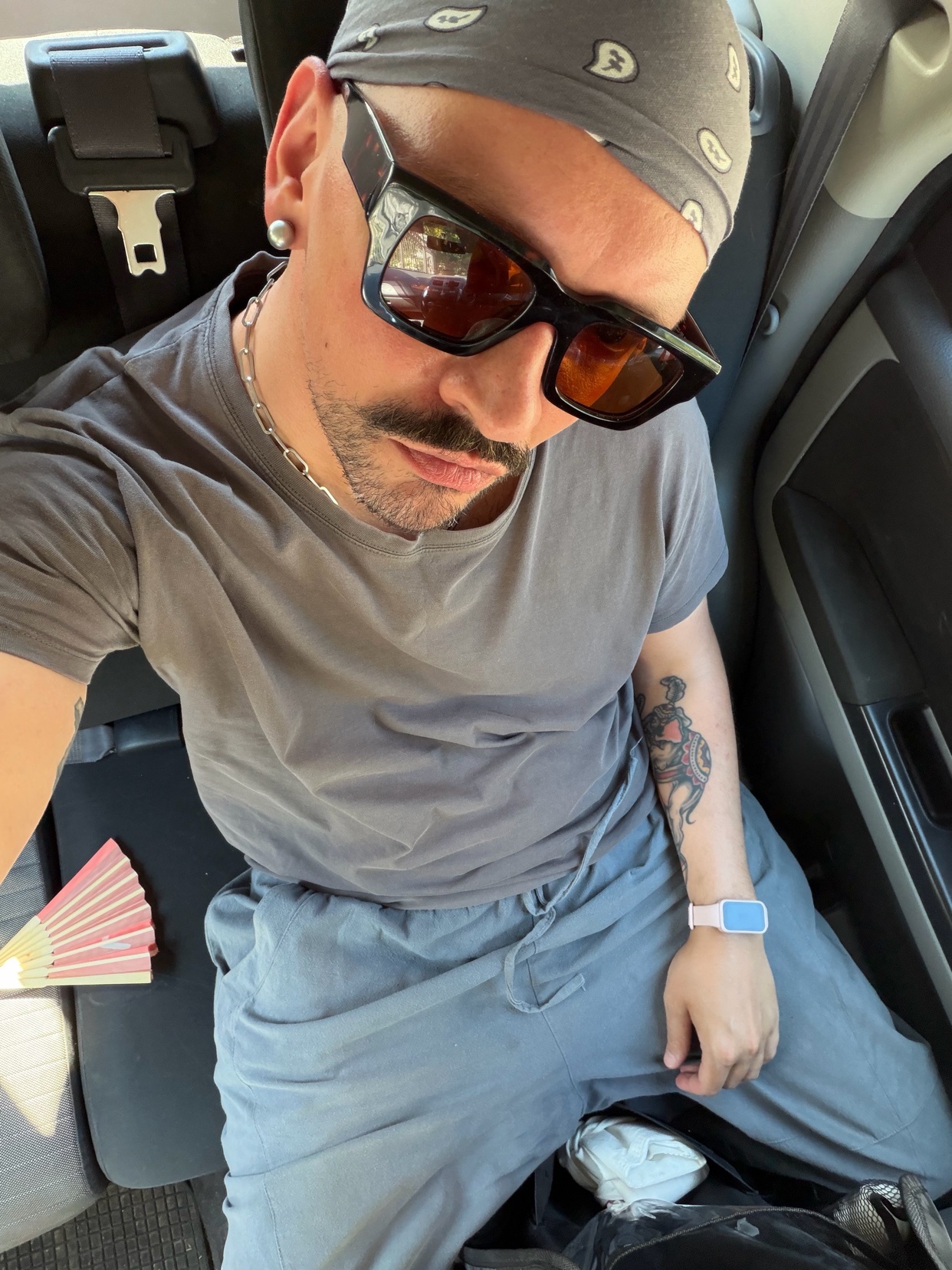 A man wearing sunglasses and a bandana poses for a selfie in a car. He has a mustache and tattoos on his arm.