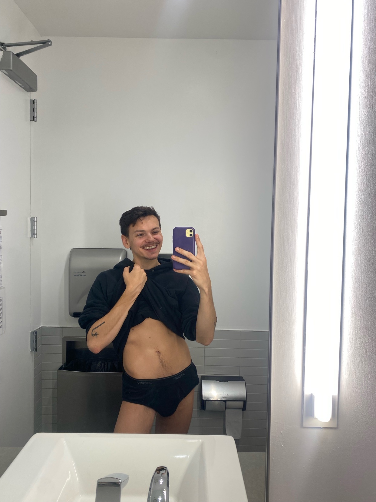 A man in black underwear and a black hoodie pulled up to his chest smiles at the camera in a bathroom selfie. He has short dark hair, a moustache, and stubble.