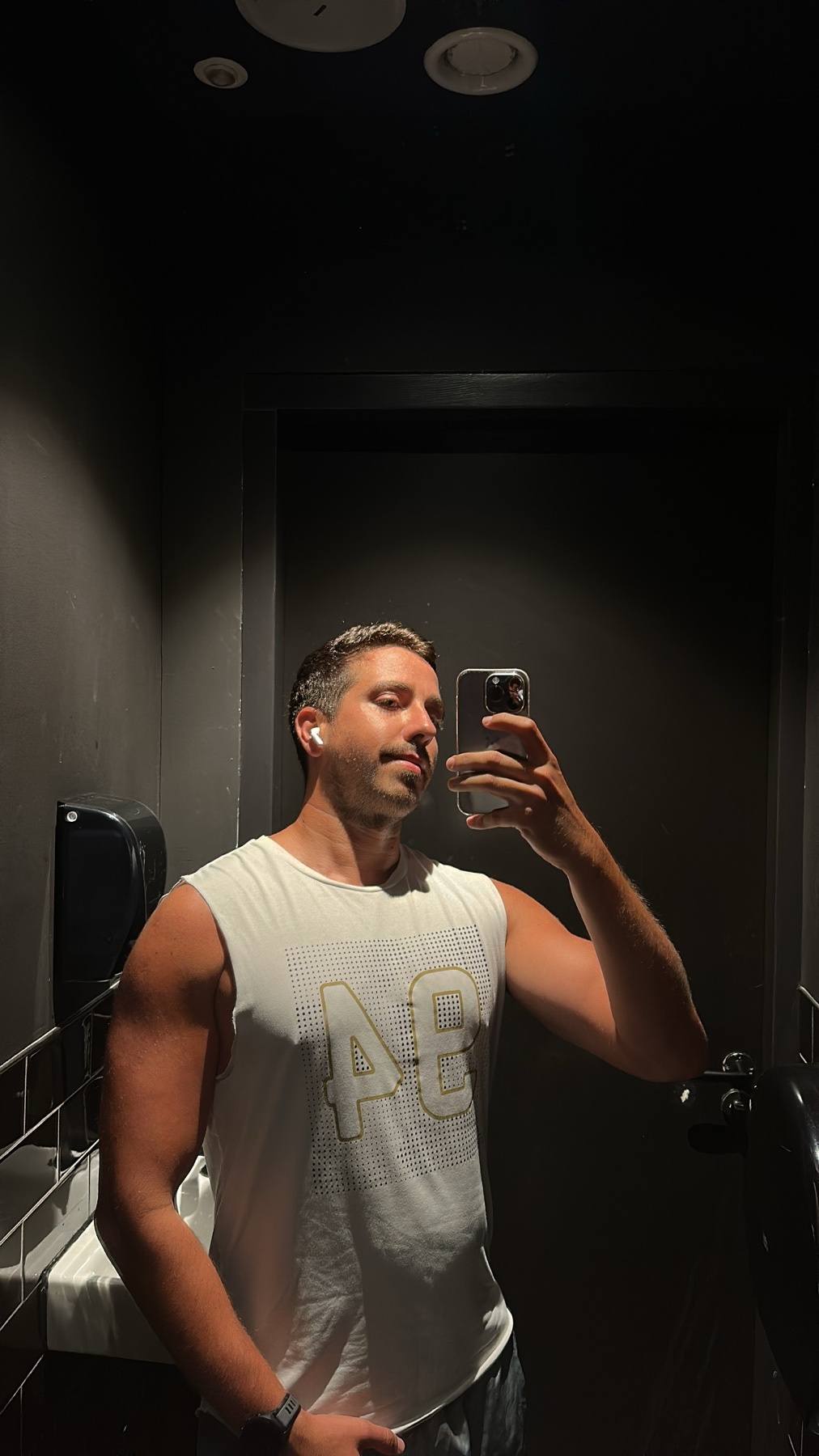 A man takes a selfie in a dimly lit bathroom, wearing a white tank top and earbuds. He has a mustache and appears to be flexing his arm.