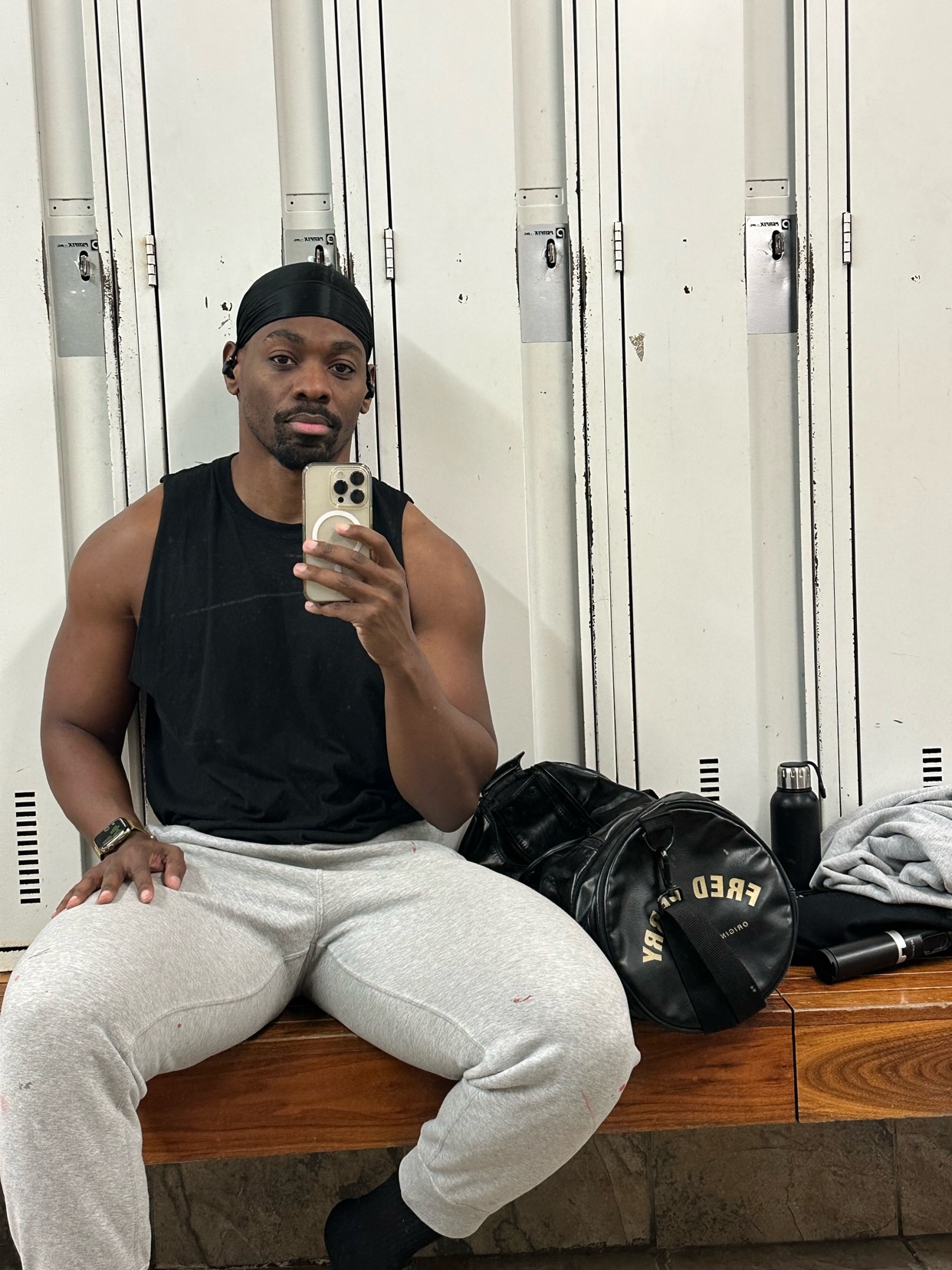 A man takes a selfie in a gym locker room. He is shirtless, showing off his muscular physique.