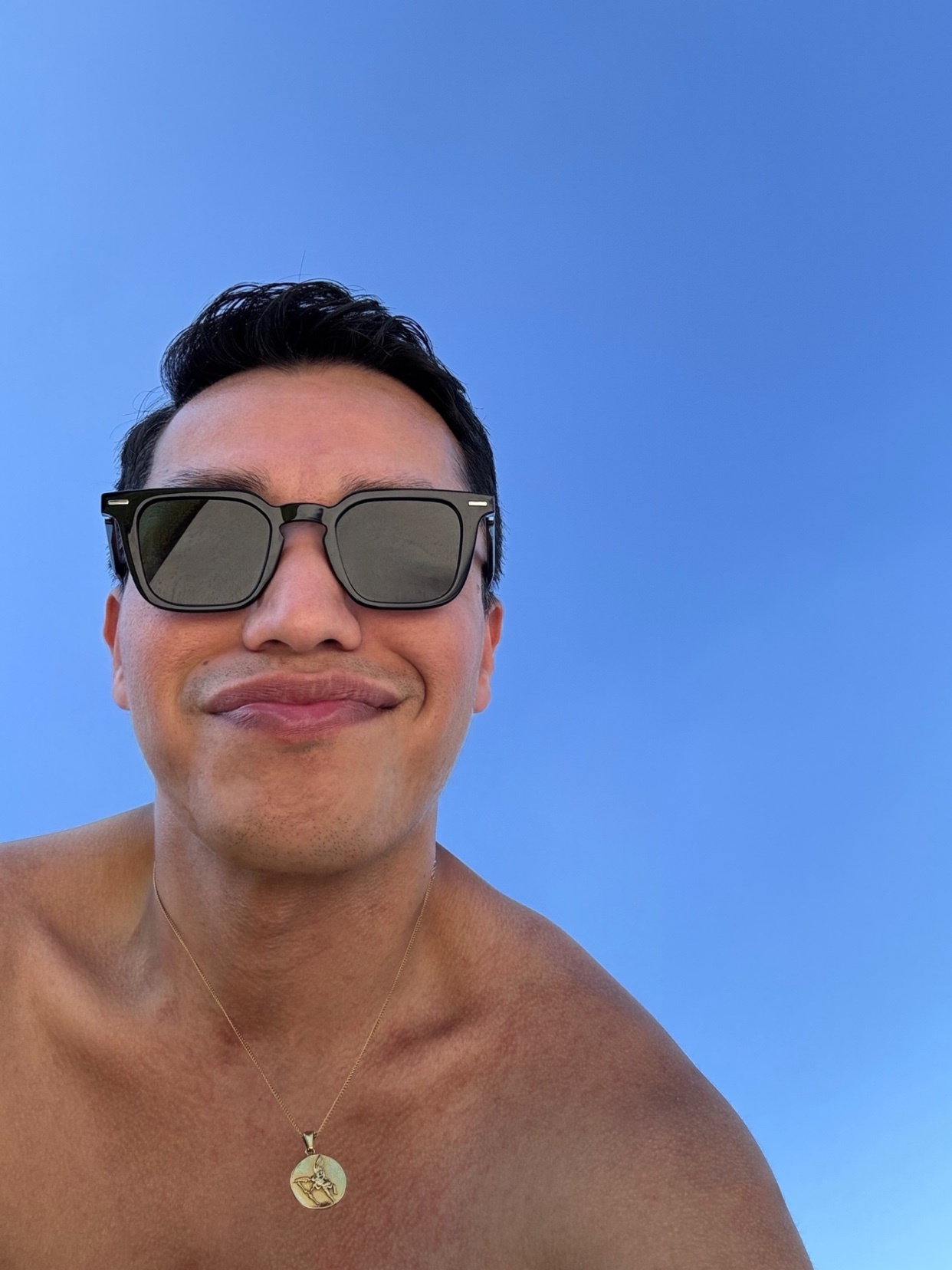 A man smiles at the camera, wearing sunglasses and a gold necklace. The photo is taken from a low angle against a bright blue sky.