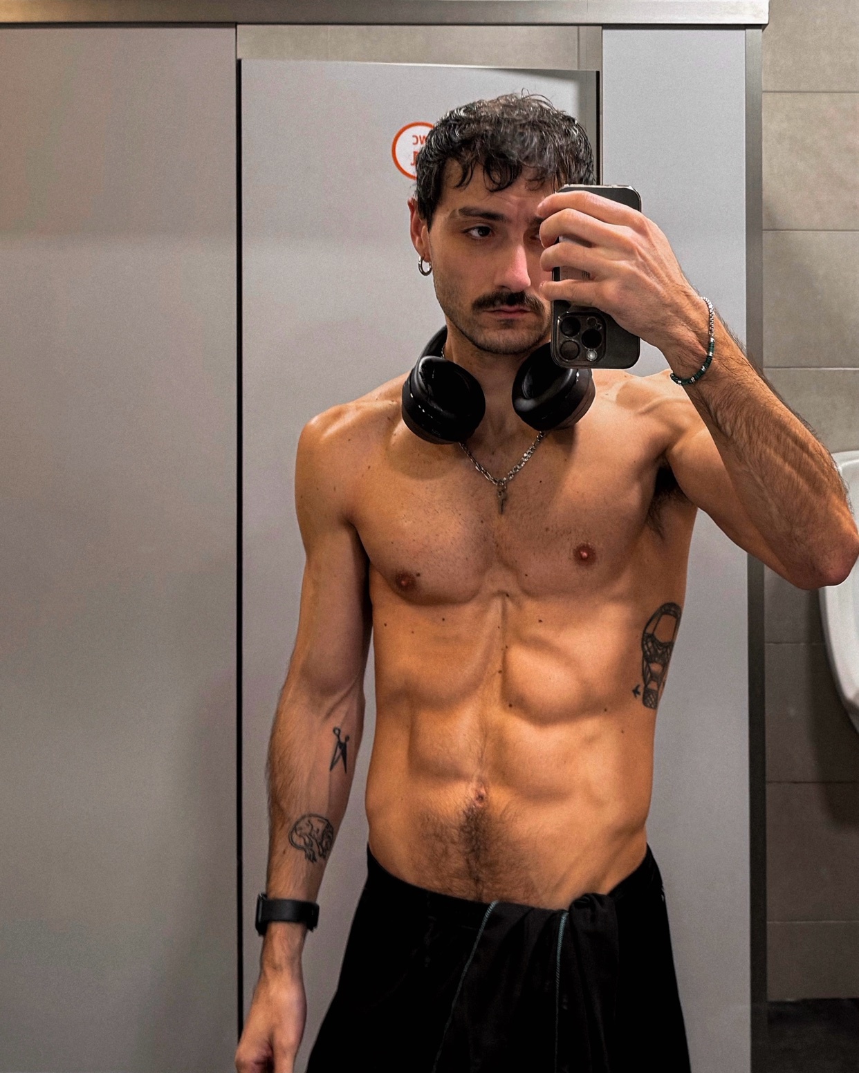A man takes a mirror selfie, showcasing his muscular physique. He is shirtless, with tattoos and headphones around his neck.