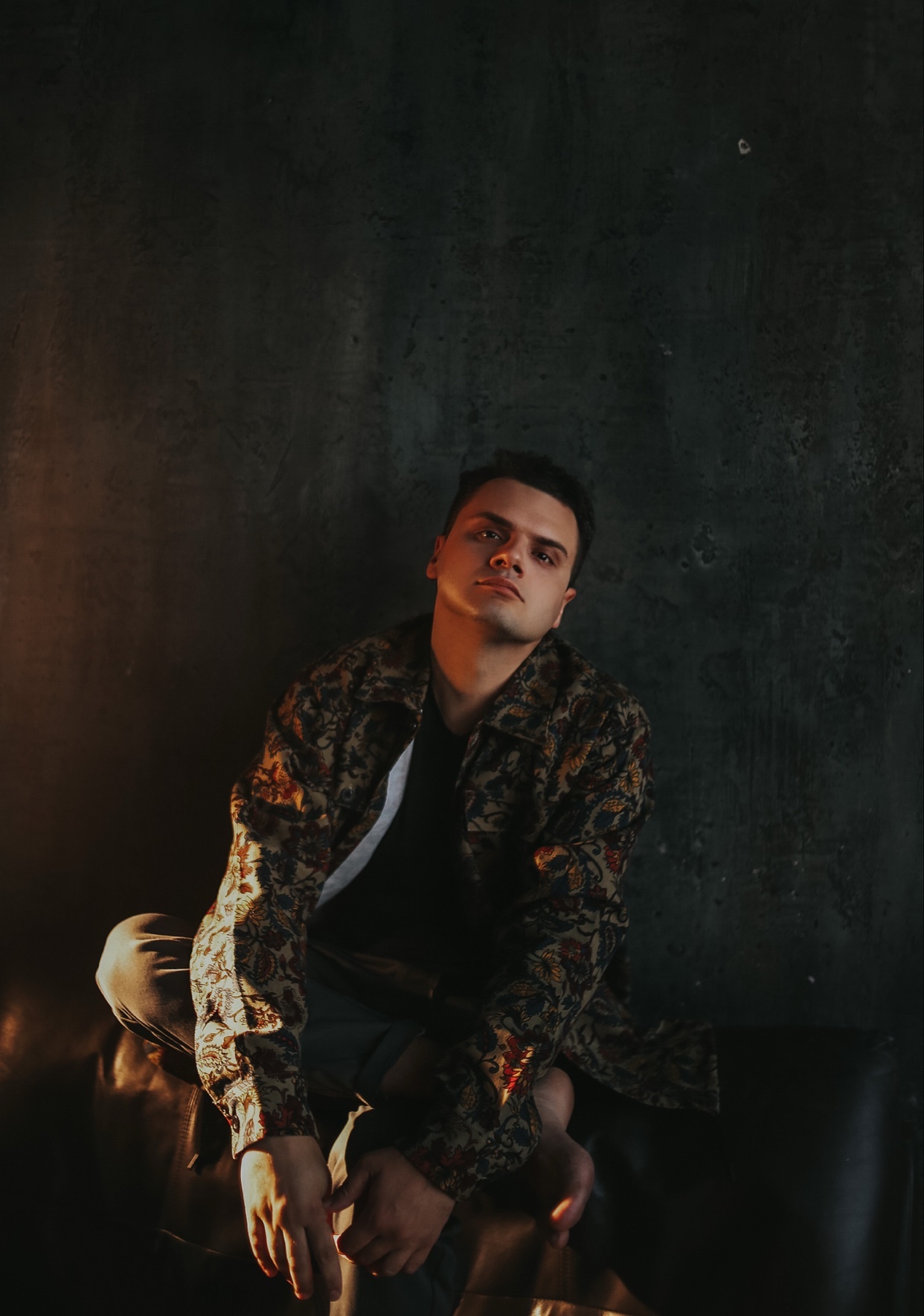 A man sits on a leather couch wearing a camouflage jacket over a dark t-shirt. The dark background and dramatic lighting create a moody portrait.