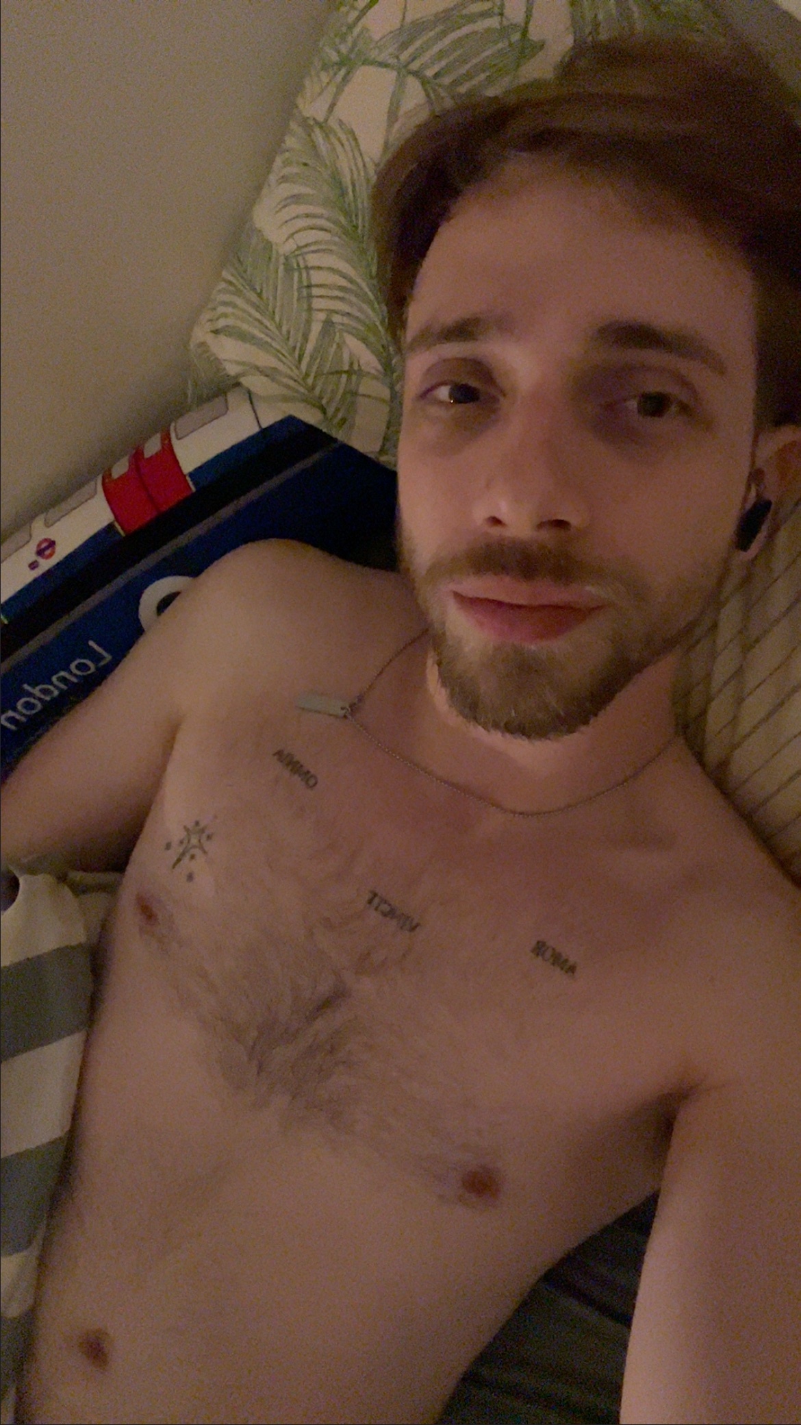 A man with a beard and tattoos lies in bed, wearing earbuds and looking at the camera.