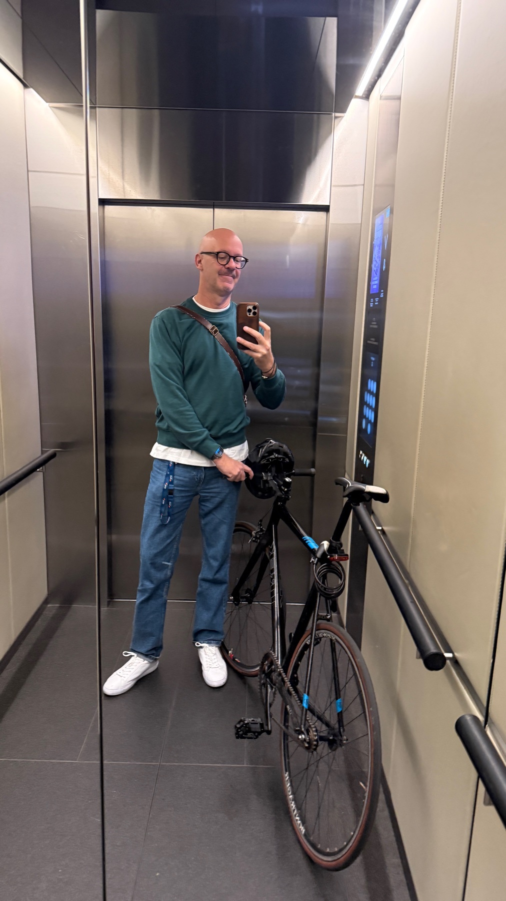 A bald man with glasses takes a selfie in an elevator with his bicycle. He is wearing a green sweatshirt and blue jeans, and appears to be returning home from work.
