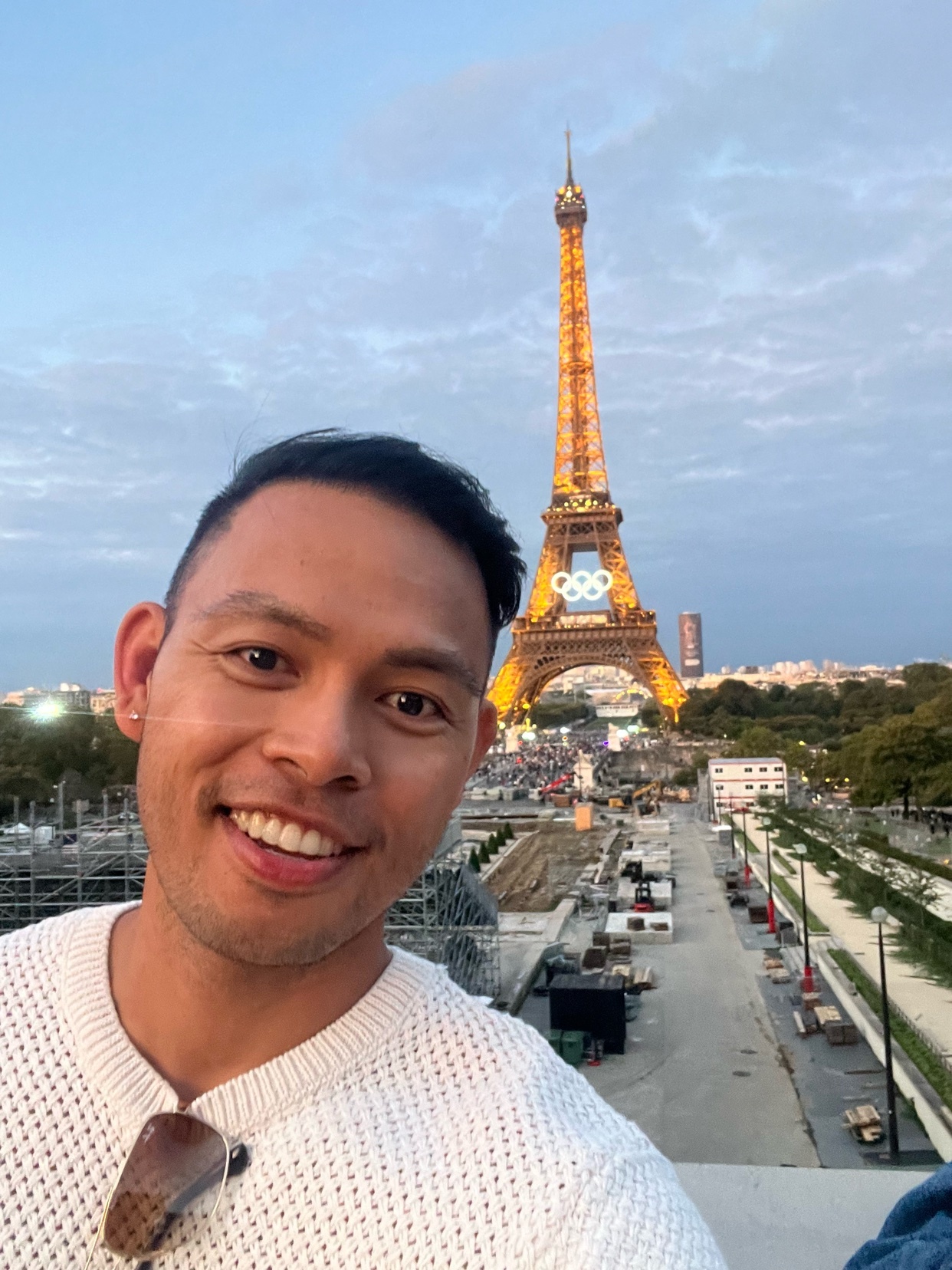 A man smiles for a selfie with the Eiffel Tower lit up in the background. The iconic Parisian landmark is adorned with Olympic rings, suggesting a connection to the upcoming games.