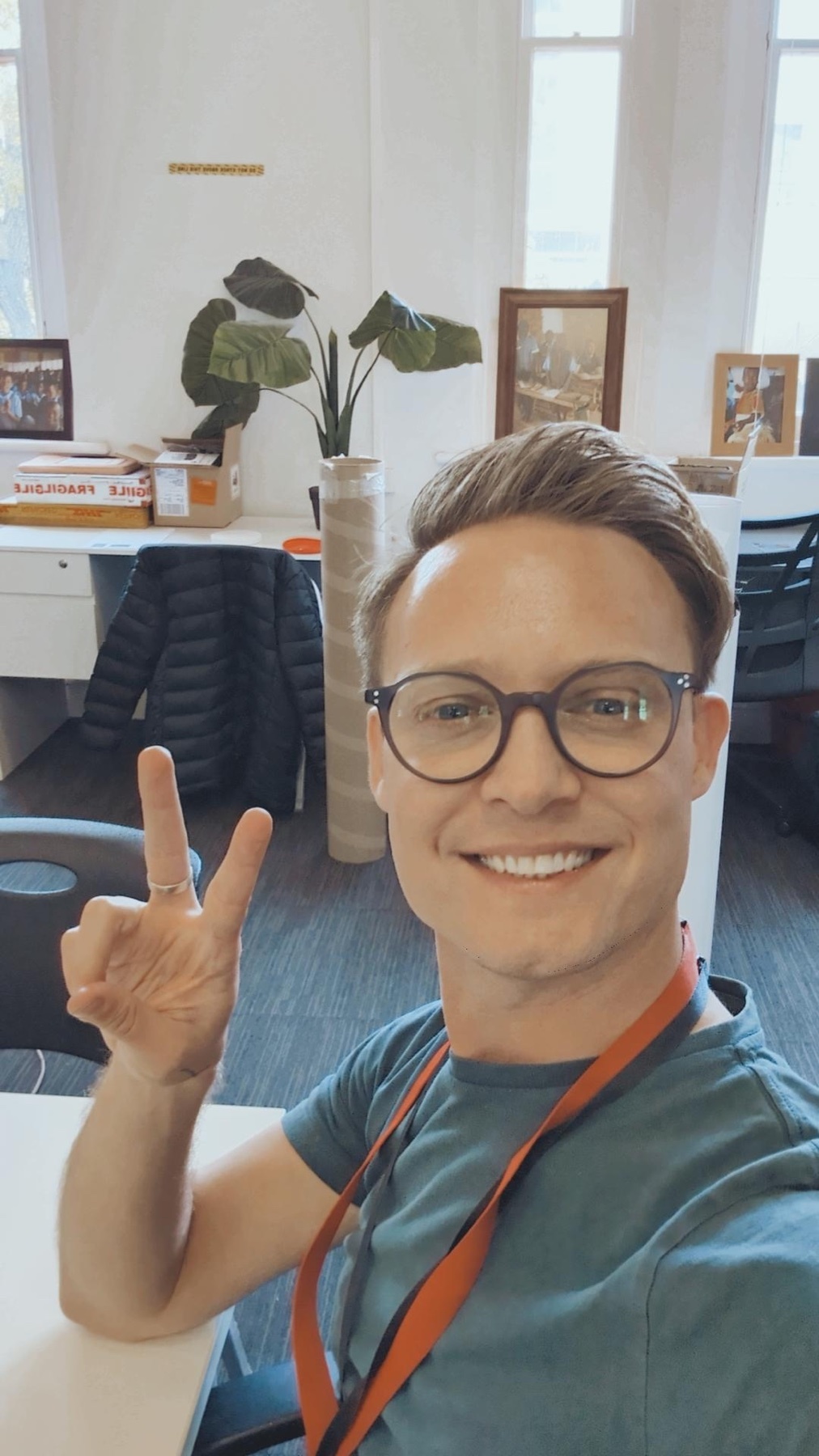A man with glasses smiles and makes a peace sign while sitting at a desk in an office setting. A large plant is visible behind him.