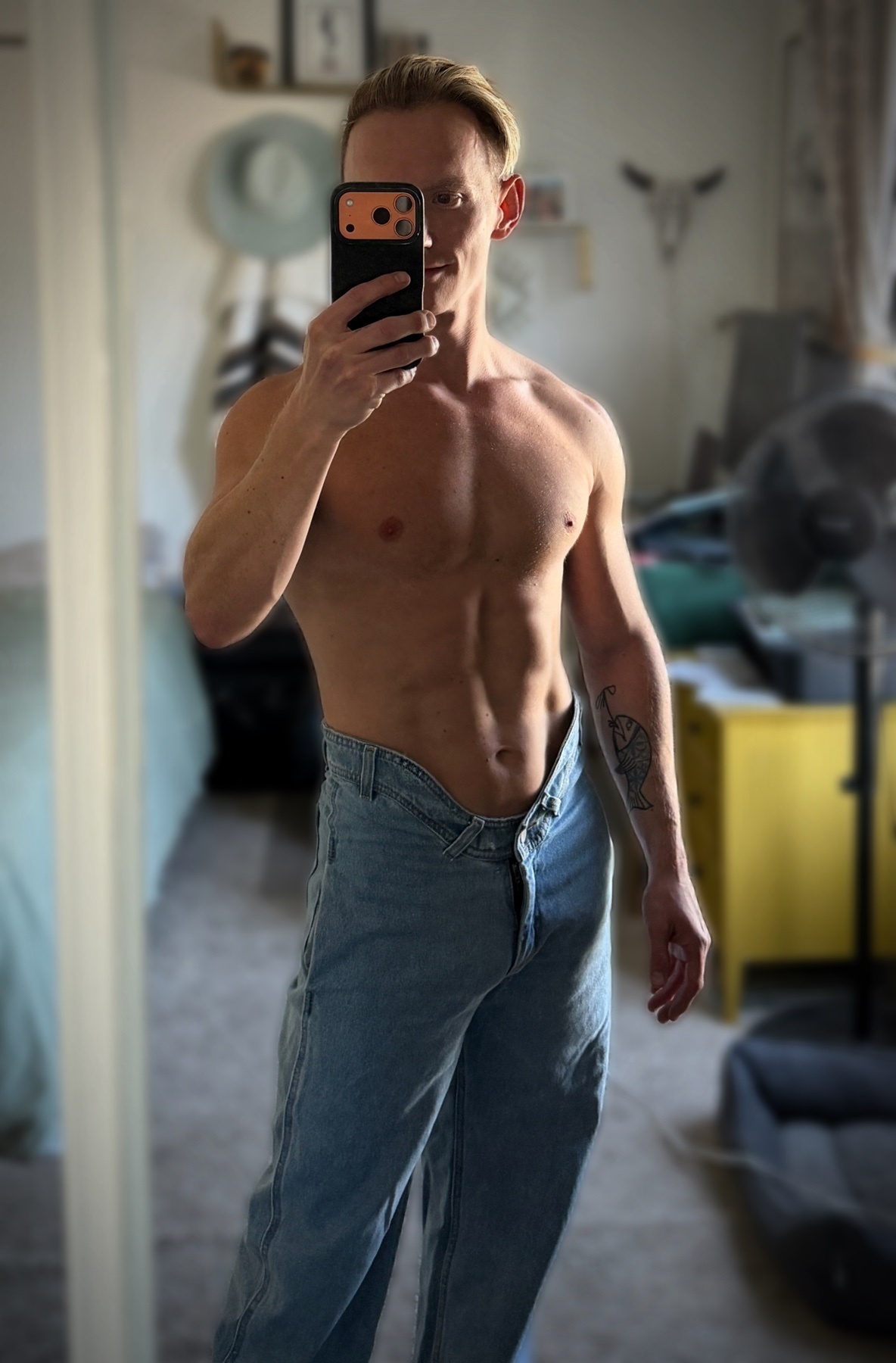 A man in light-wash jeans takes a mirror selfie in a room. He is shirtless, showing off his muscular physique and a tattoo on his arm.