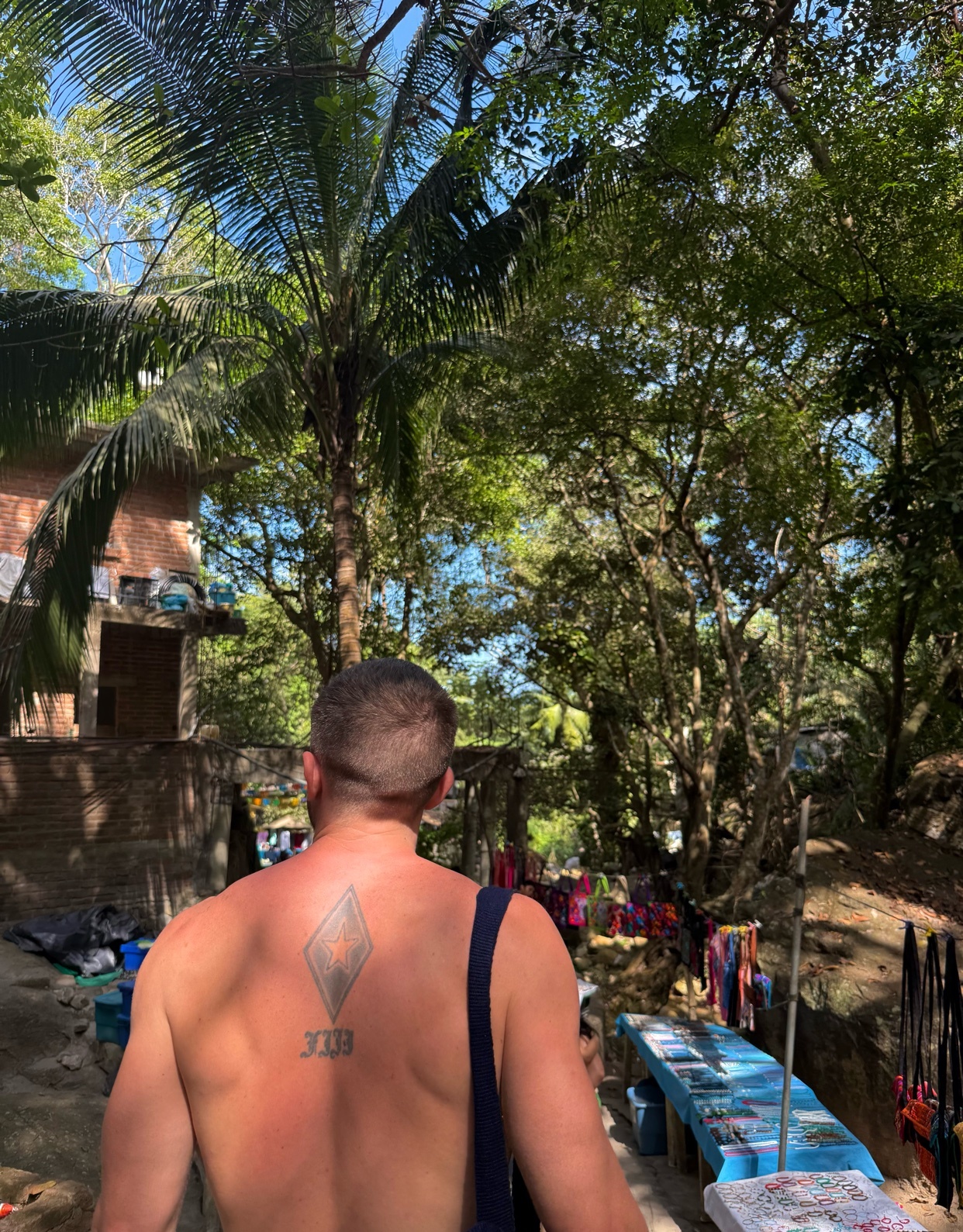 A man with a diamond tattoo on his back walks through a tropical outdoor setting. The photo is taken from behind, showcasing the man's bare back and the lush greenery.