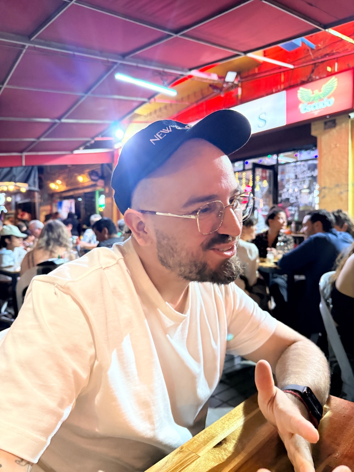 A man wearing glasses and a cap is sitting at a restaurant. The photo is taken at night.