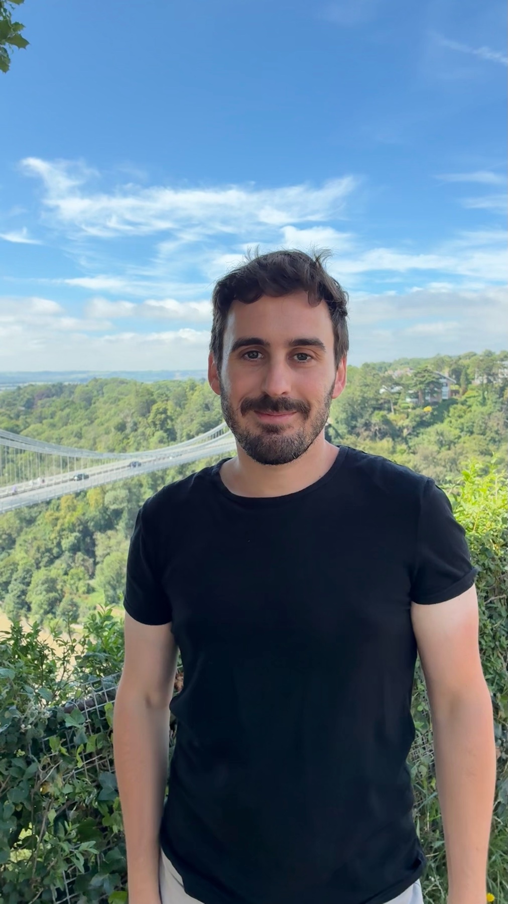 A man with dark hair and stubble stands outdoors in front of a scenic view with a bridge in the background. He is wearing a black t-shirt and appears to be casually posing for the photo.
