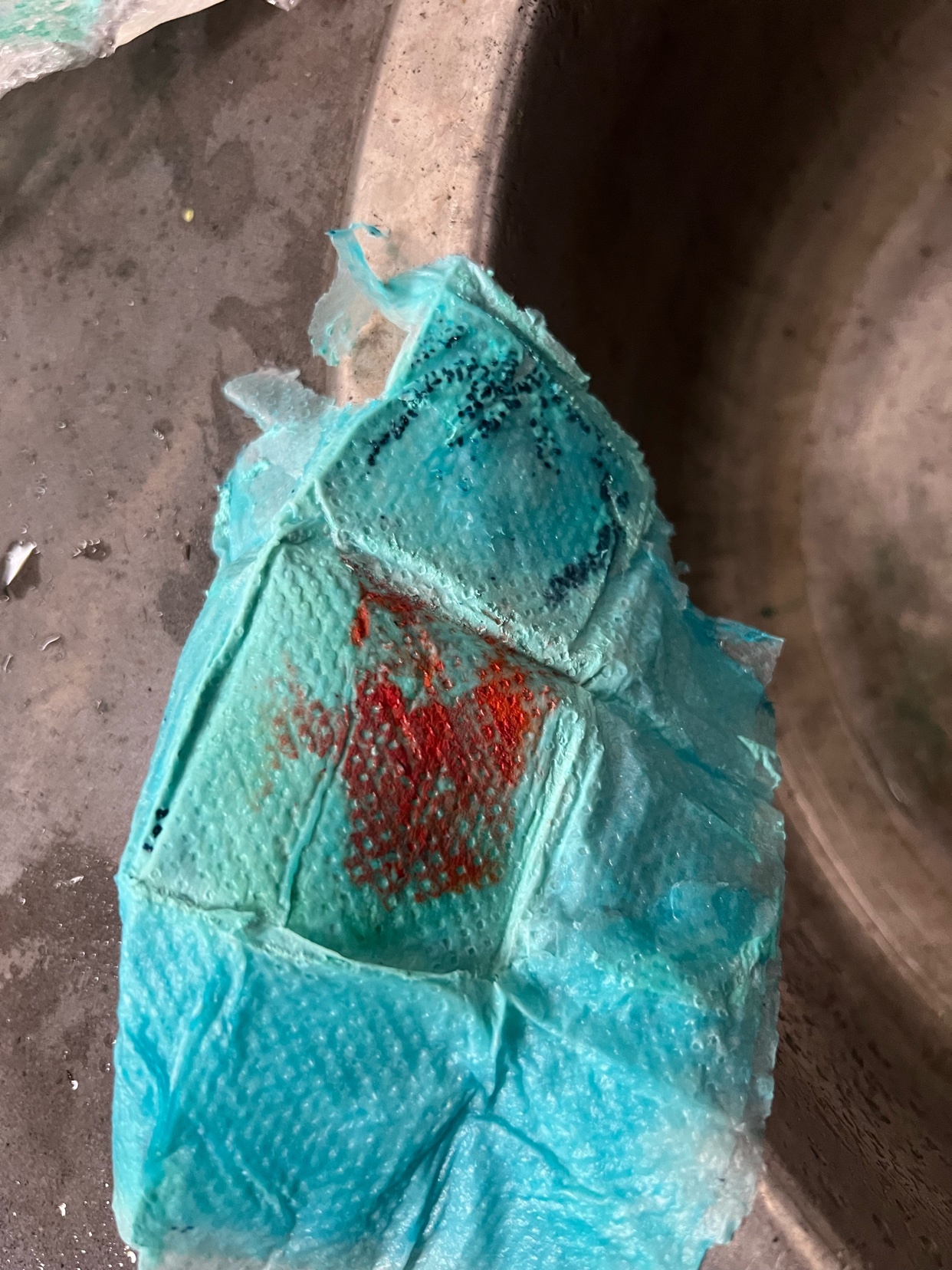 A close-up shot of a crumpled blue paper towel with red and black markings, likely from a chemical reaction creating a copper patina. The texture of the paper towel is visible, highlighting the artistic process of metal finishing.
