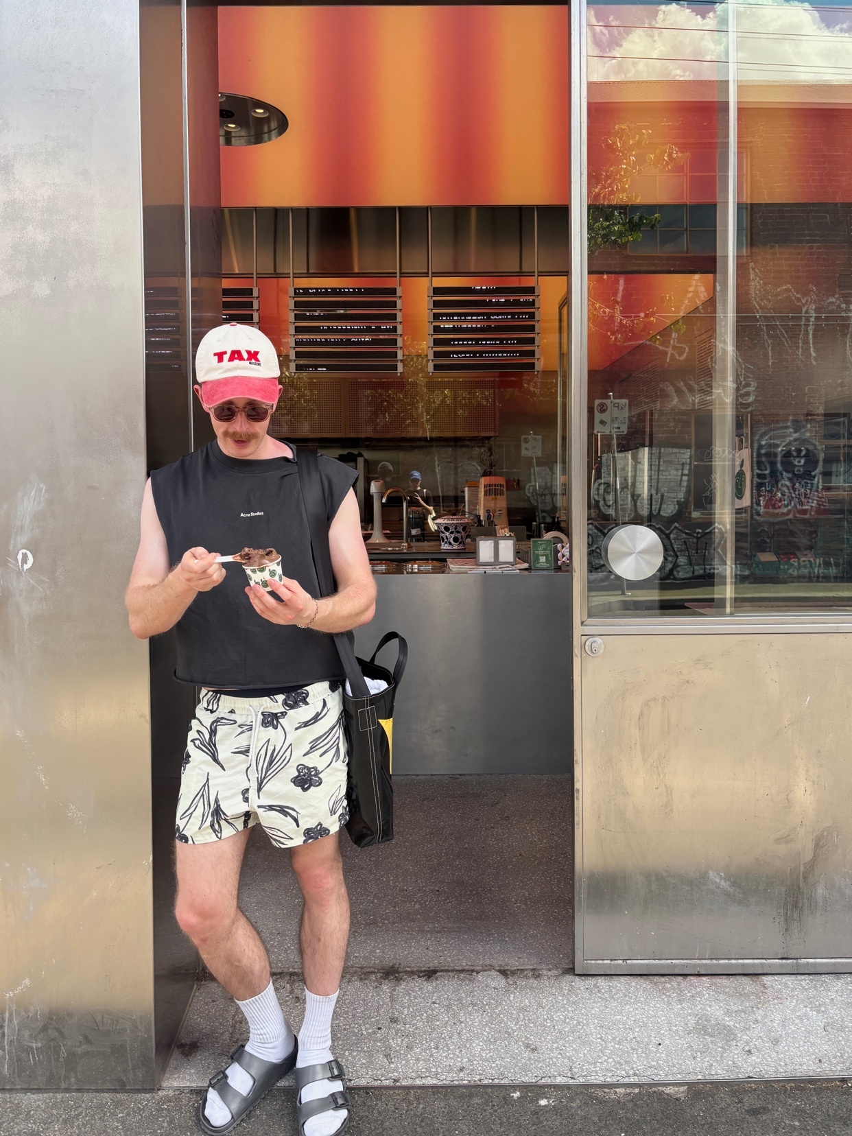 A man in a black sleeveless shirt and floral shorts enjoys ice cream. He is wearing a cap and sunglasses, standing in front of a shop.