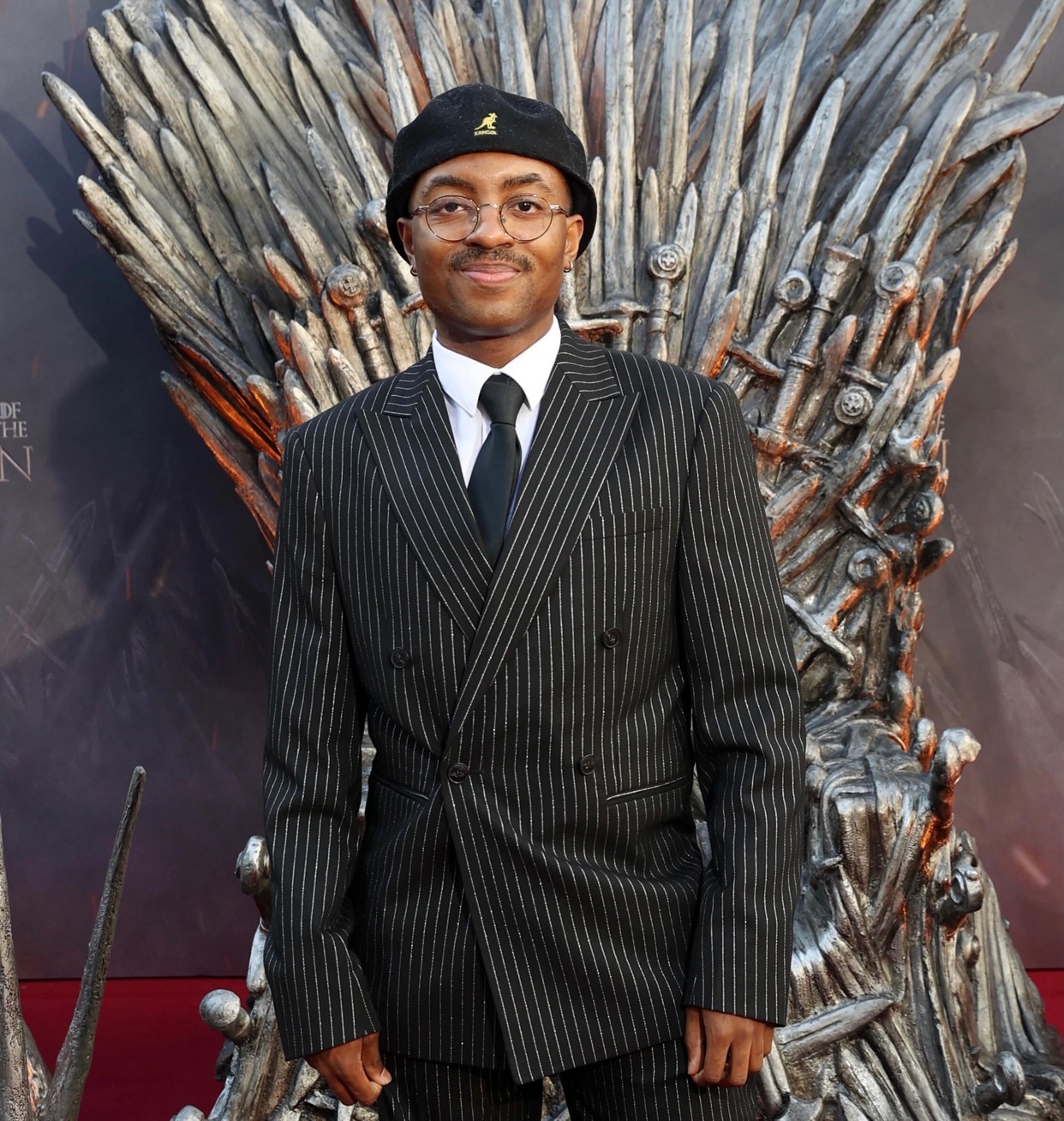 A man in a pinstripe suit and black hat poses in front of the Iron Throne.