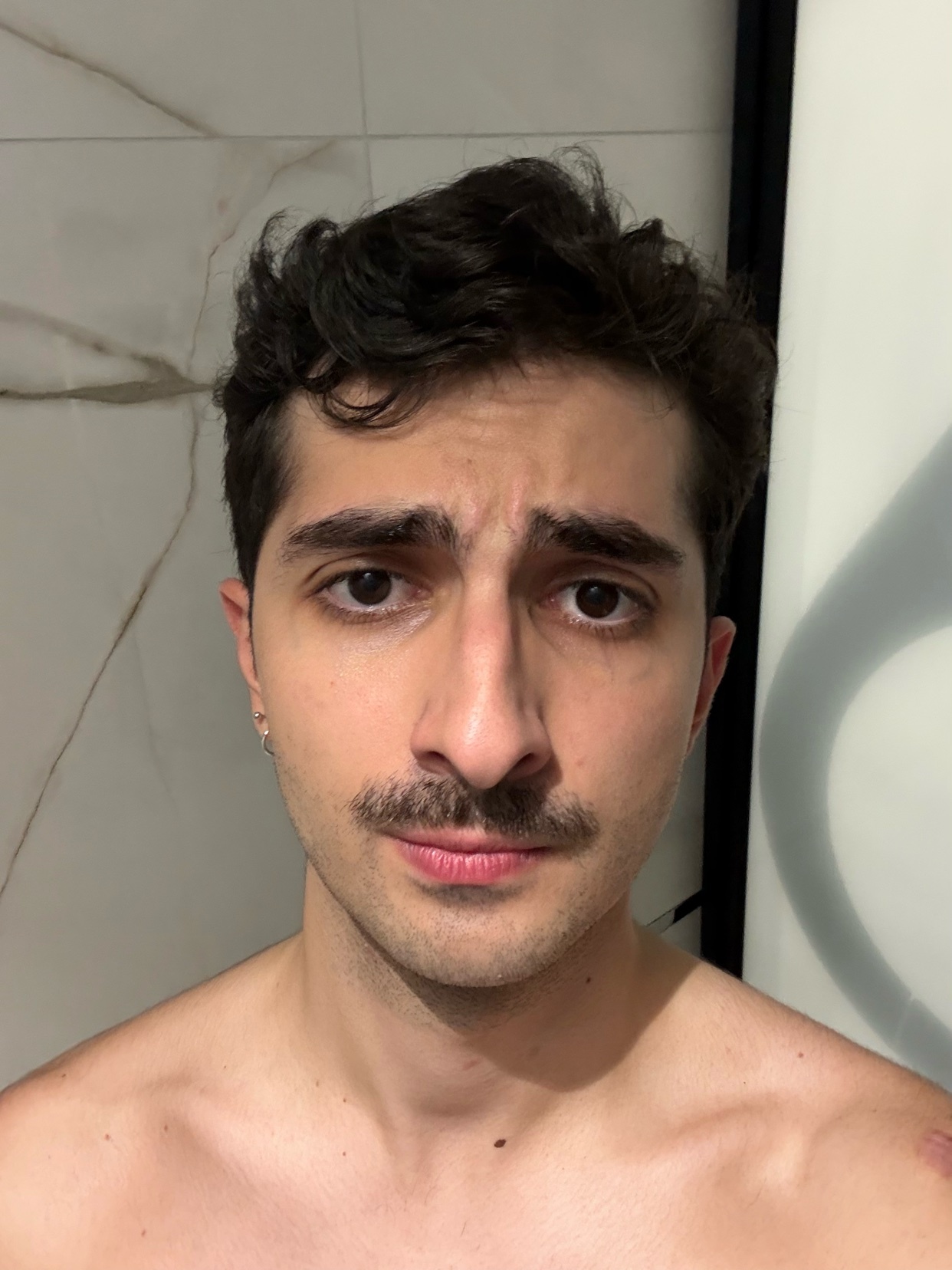 A man with dark, curly hair and a mustache takes a selfie. He is shirtless.