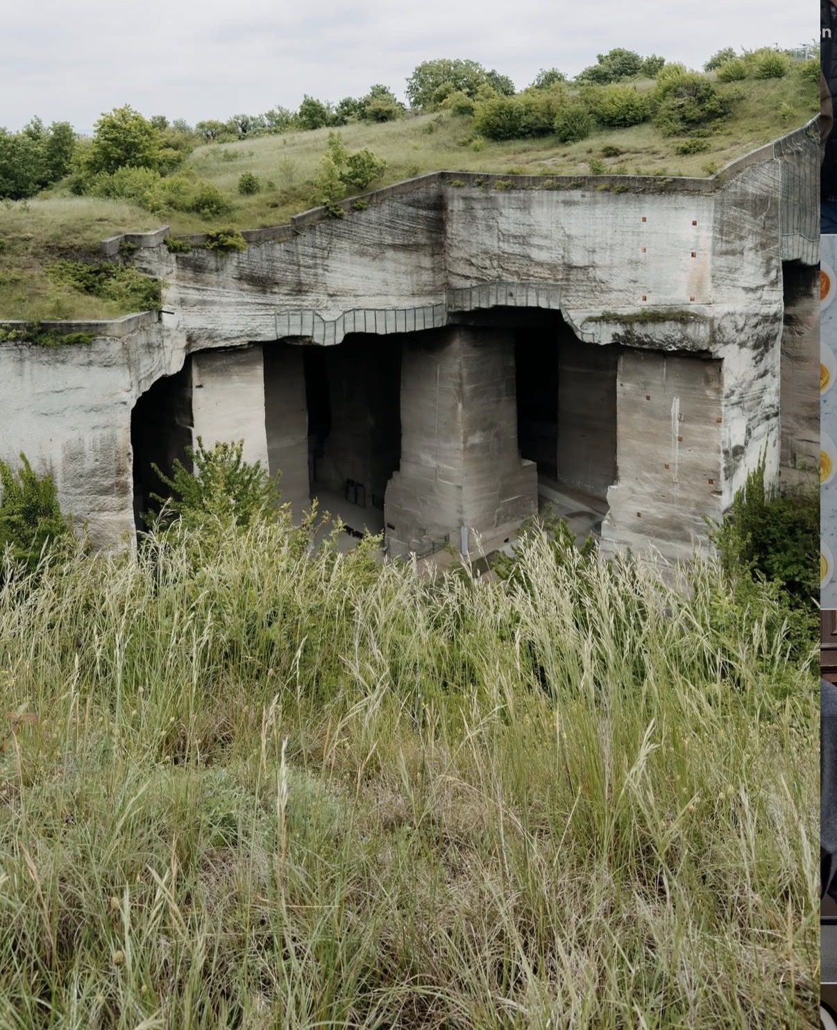 An overgrown quarry with large stone pillars and structures carved into the earth. Tall grasses and green foliage surround the abandoned industrial site.