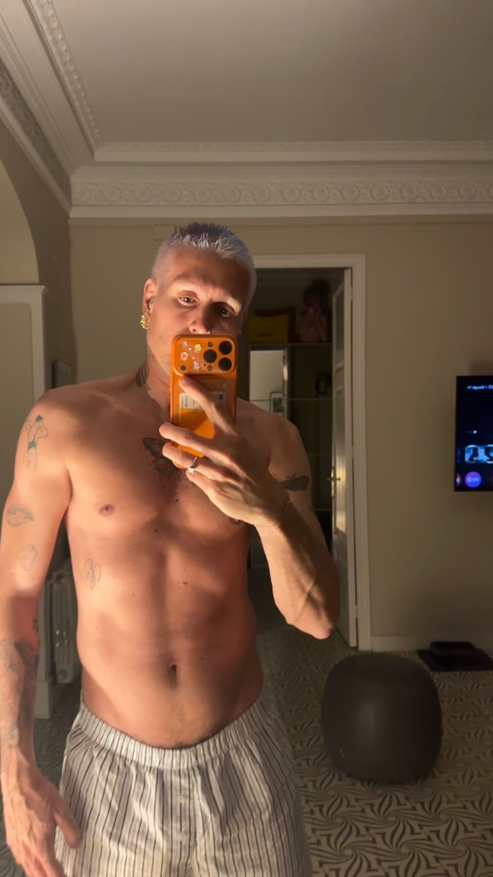 A man with blue hair and tattoos takes a mirror selfie while wearing pajama pants. He is shirtless, showing off his muscular physique.