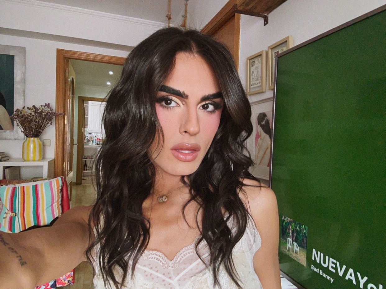 A woman with long, dark, wavy hair and dramatic makeup poses for a selfie in front of a television screen.