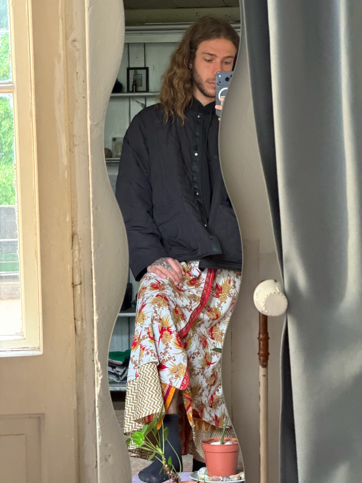 A man with long, wavy brown hair takes a mirror selfie in a room with eclectic decor. He is wearing a black quilted jacket over a patterned skirt and black socks, holding a phone in his hand.