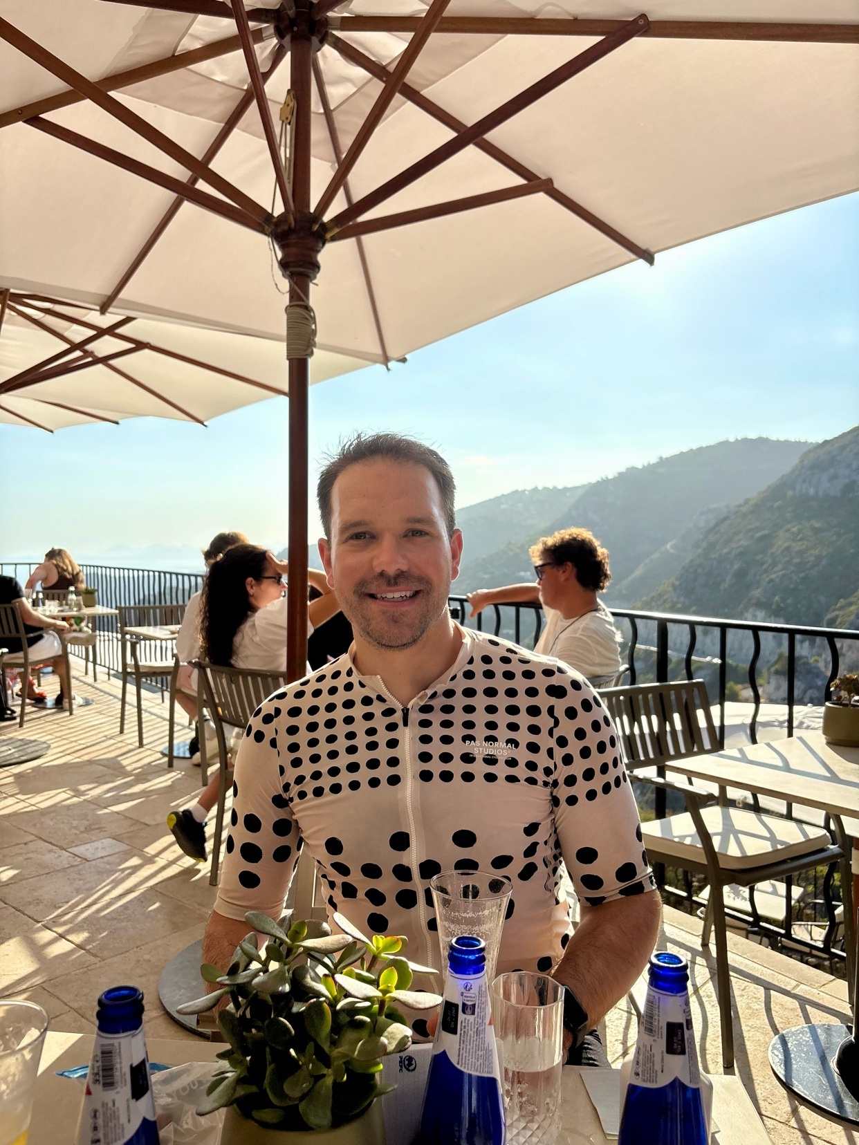 A man smiles at the camera while sitting at an outdoor table at a restaurant with a scenic mountain view.