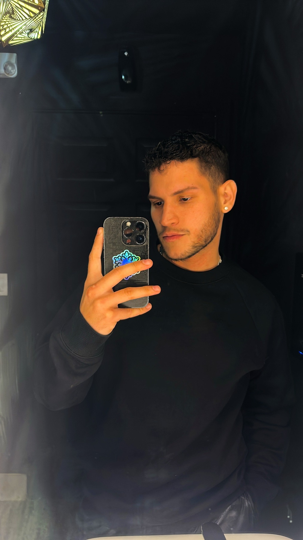 A man with dark, curly hair and stubble takes a selfie in a mirror. He is wearing a black sweatshirt and an earring.