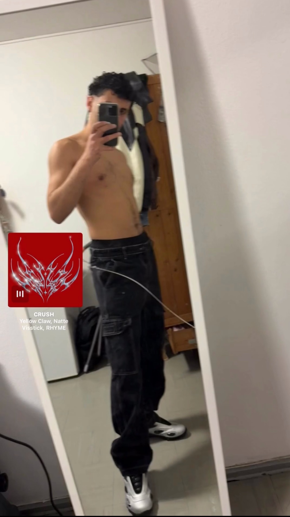A man takes a mirror selfie, shirtless, showing off his physique. He's wearing black cargo pants and sneakers.